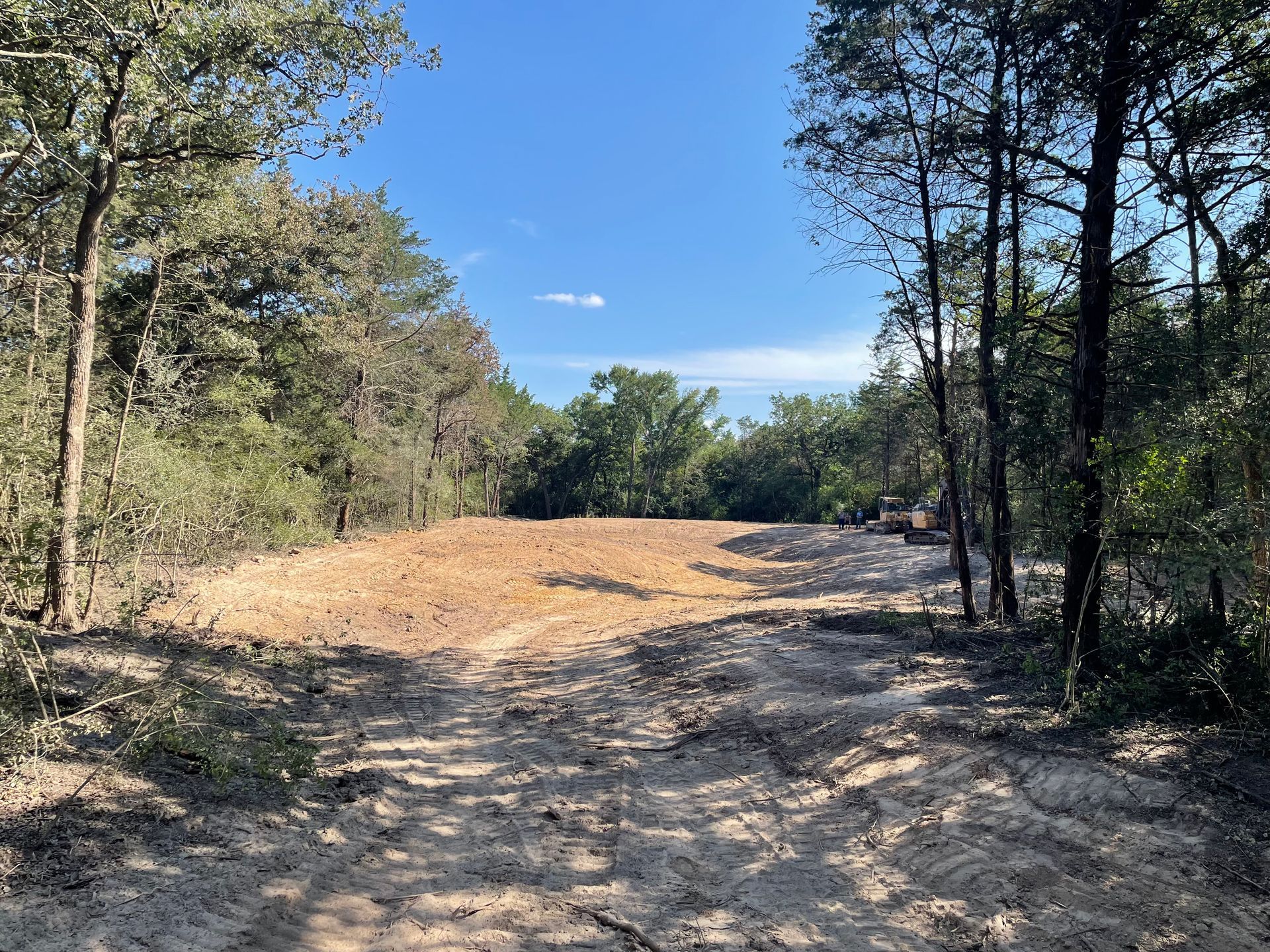 Cleared land in a forest, ready for construction. Light brown dirt, green trees, and blue sky.