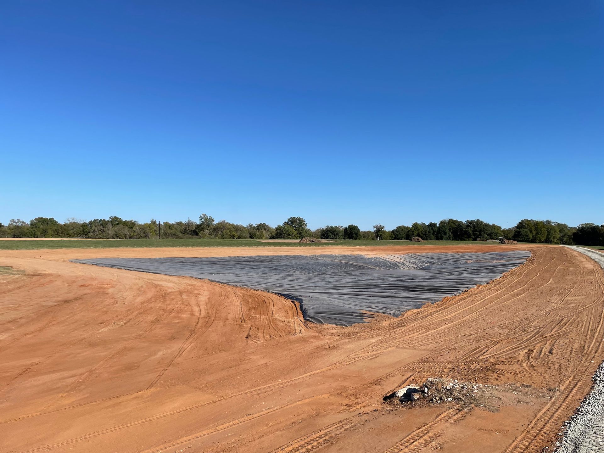 A large, earthen construction site with a water-filled, black-lined basin under a blue sky.