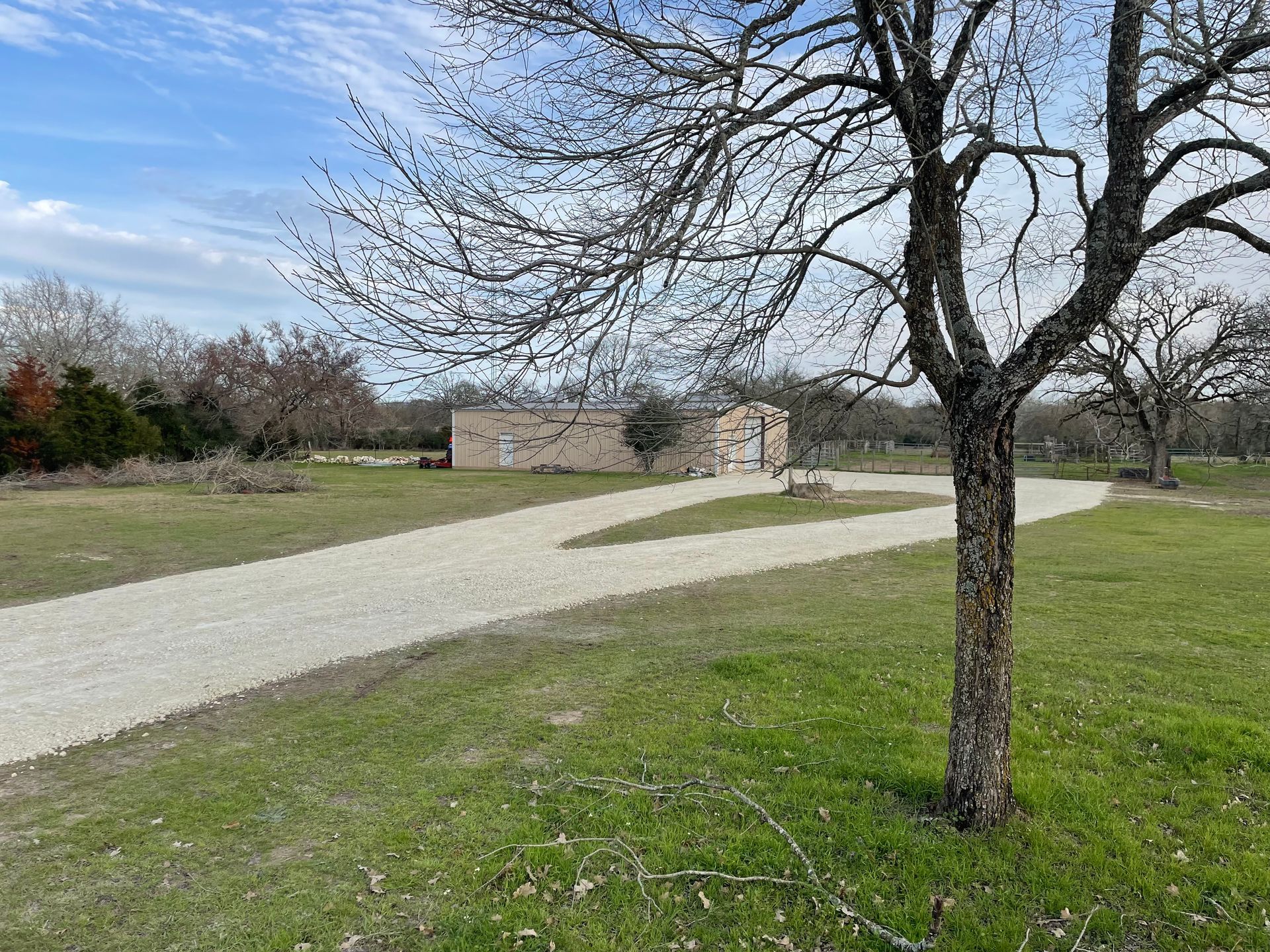 Gravel driveway leading to a tan building, a bare tree in the foreground, green grass, and a blue sky.
