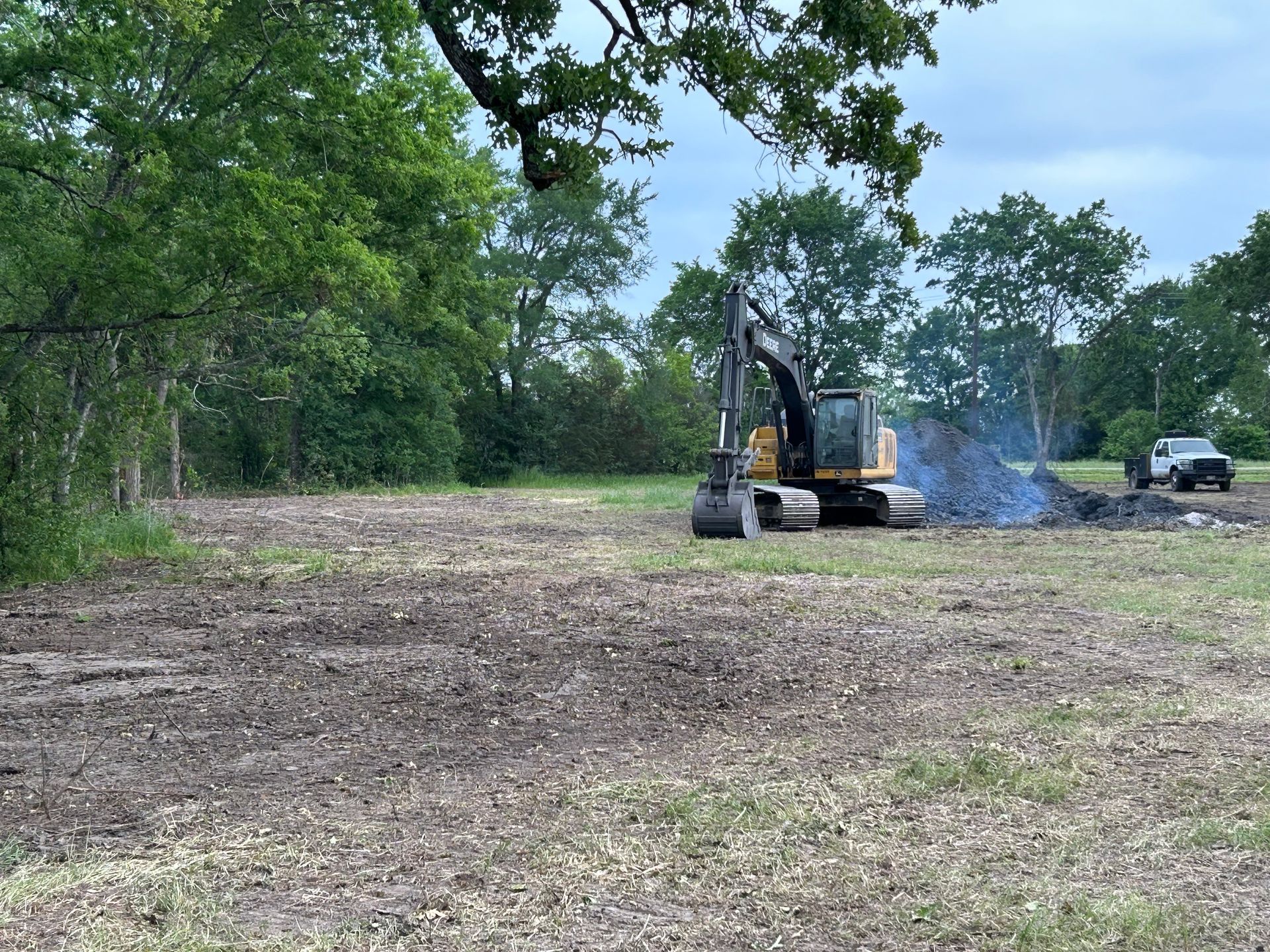 An excavator clearing a field, burning debris, with a truck nearby on a cloudy day.