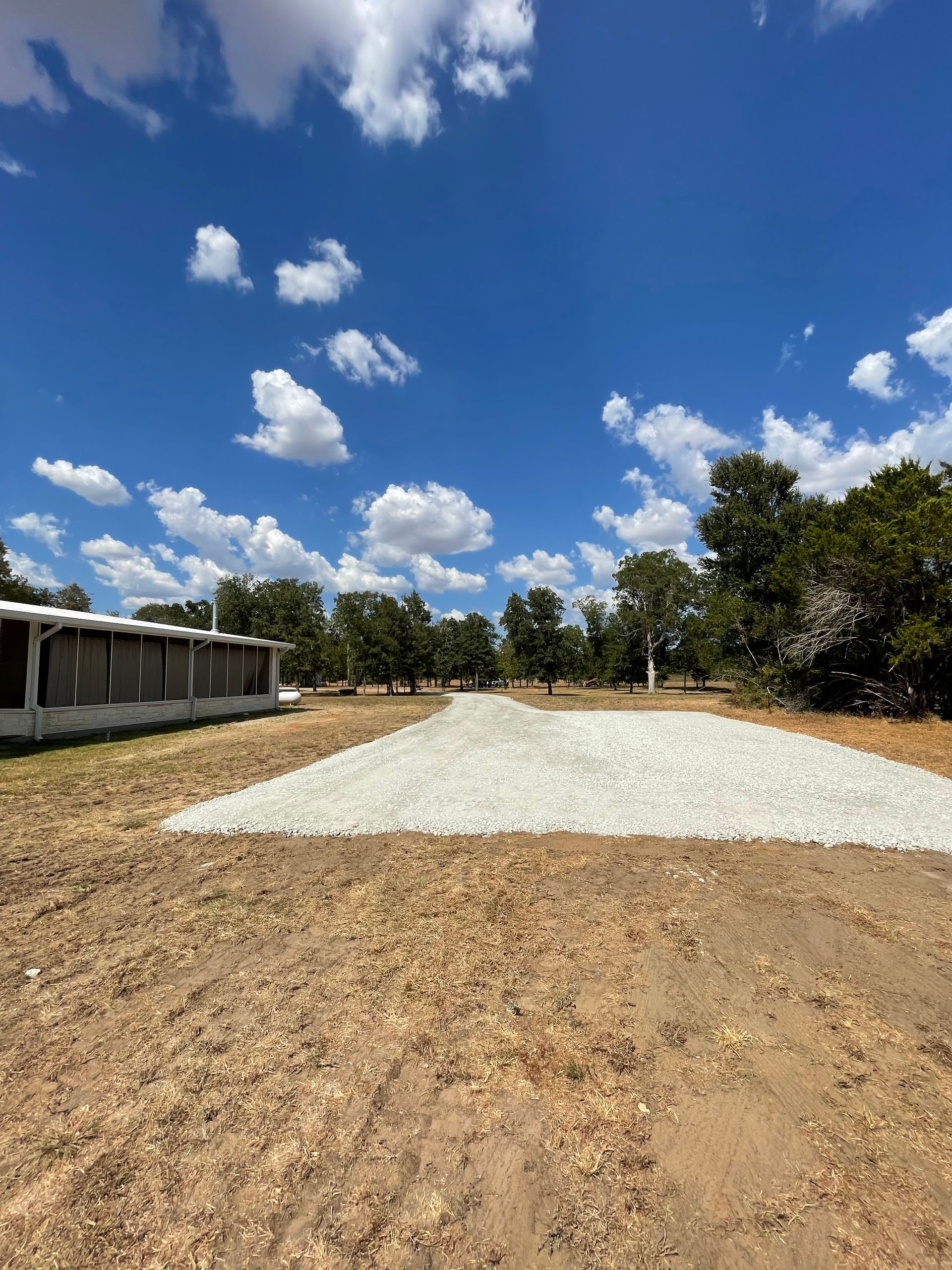 A gravel driveway leads to a treeline on a sunny day, with a blue sky and scattered clouds.