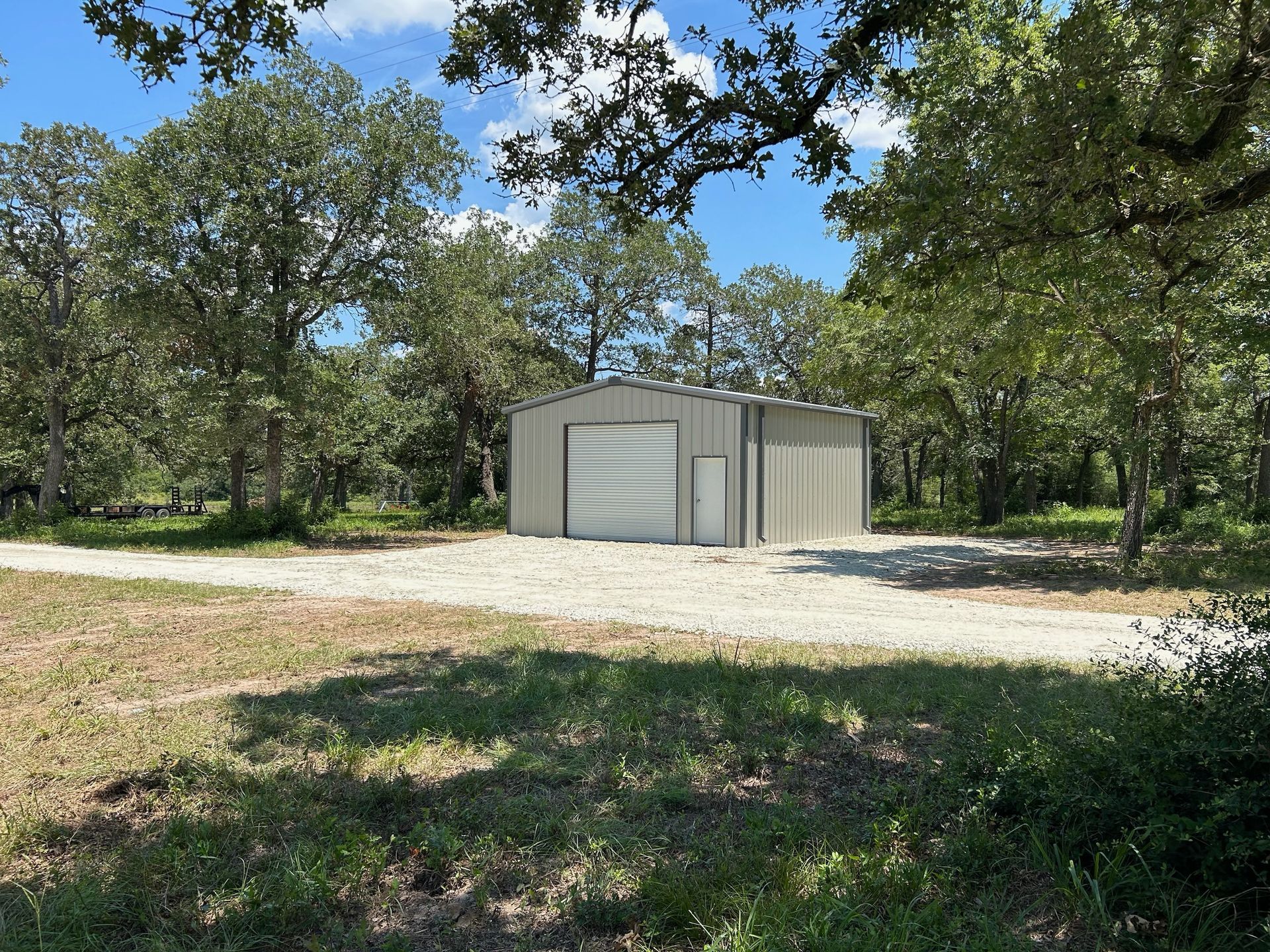 Gray metal shed on gravel driveway, surrounded by trees under a blue sky.