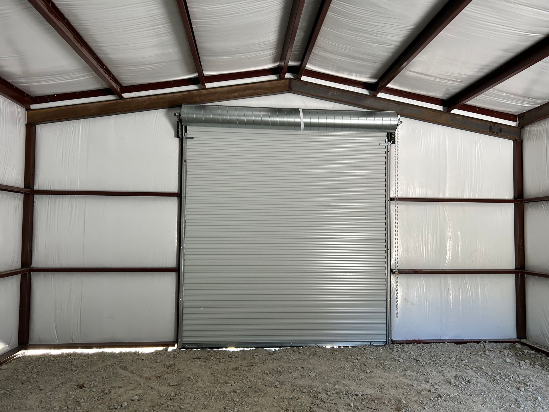 Interior view of a metal building with a closed roll-up door. White walls, brown metal beams, gravel floor.