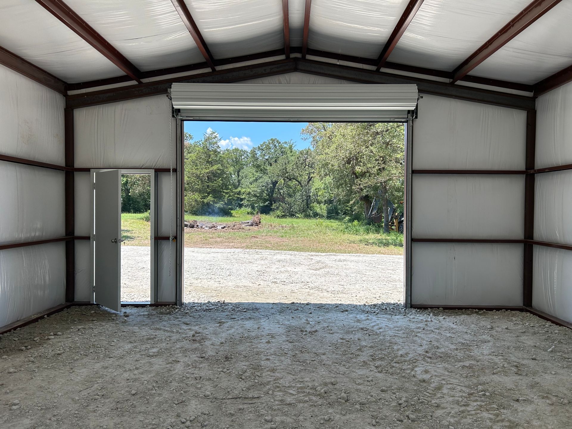 Interior of a metal building with roll-up door open to a gravel area and trees. Small door visible.