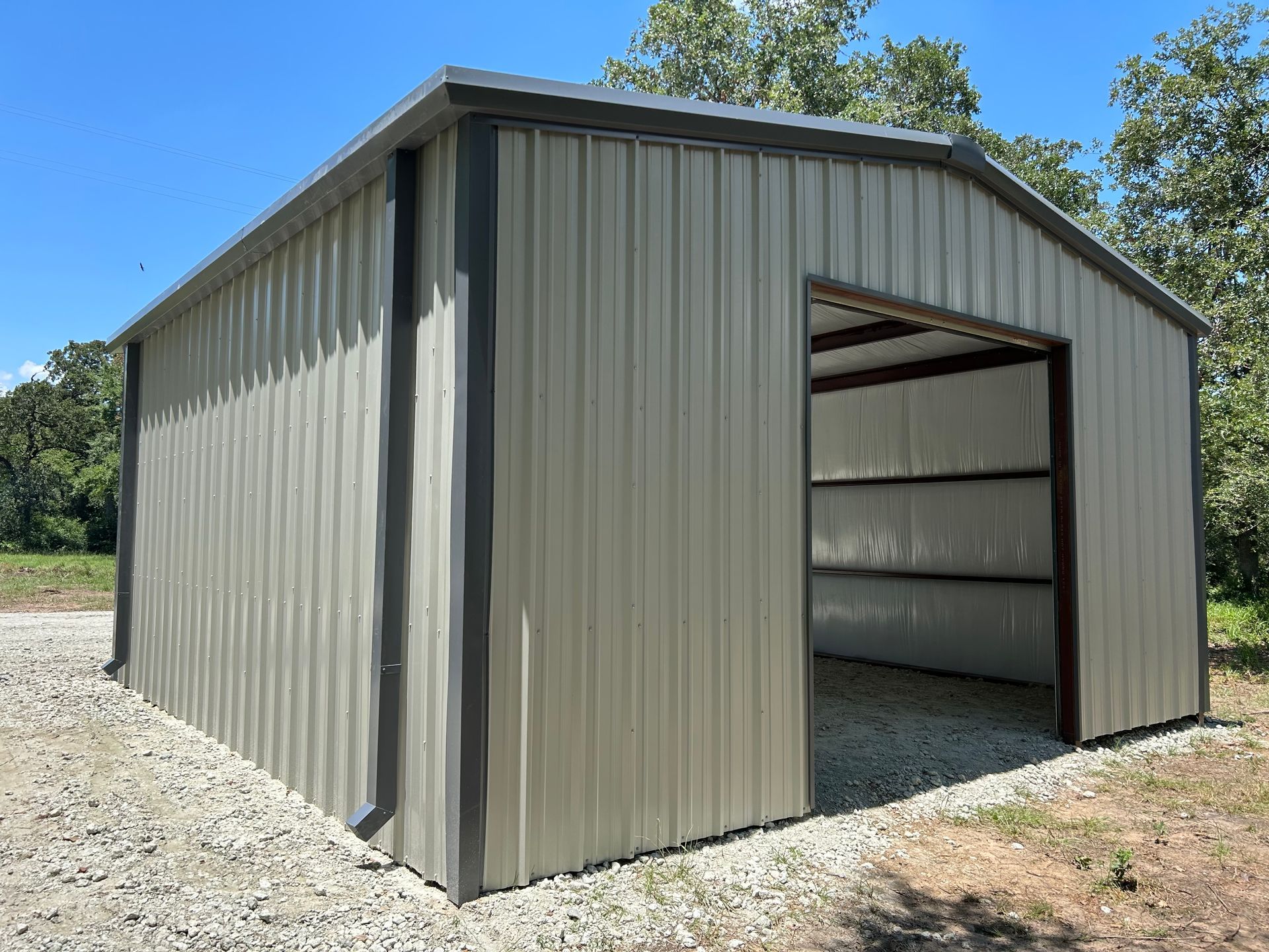 Tan metal storage shed with dark trim and open doorway on gravel.