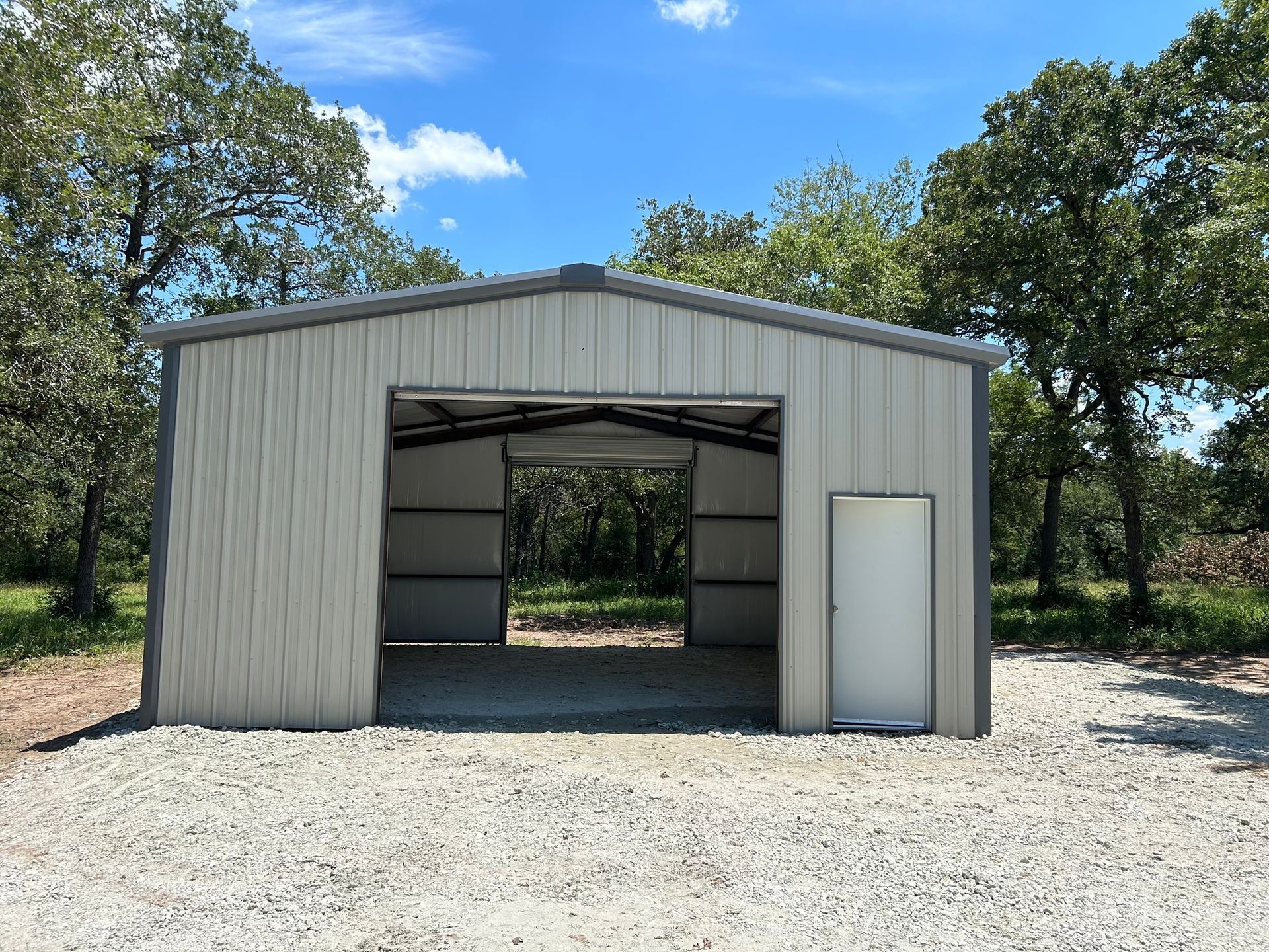 Metal building with open garage door and white entry door on a gravel surface, surrounded by trees under a blue sky.