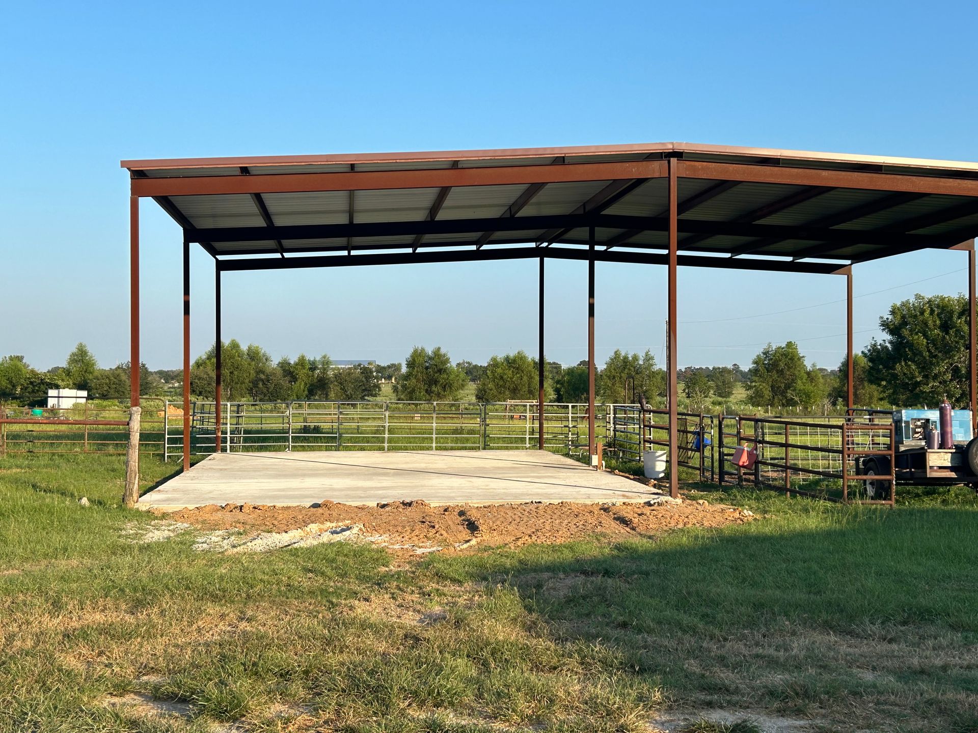 A metal-framed shelter with a concrete base on a grassy field, with surrounding fences and trees in the background.