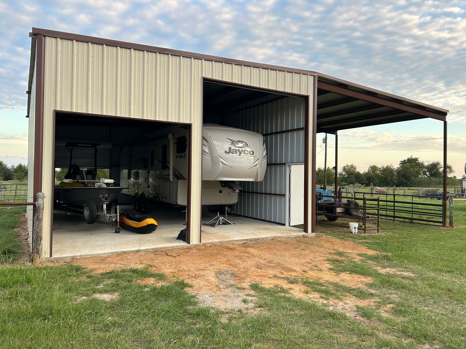 RV parked under a metal carport with a concrete pad, in a grassy field under a cloudy sky.