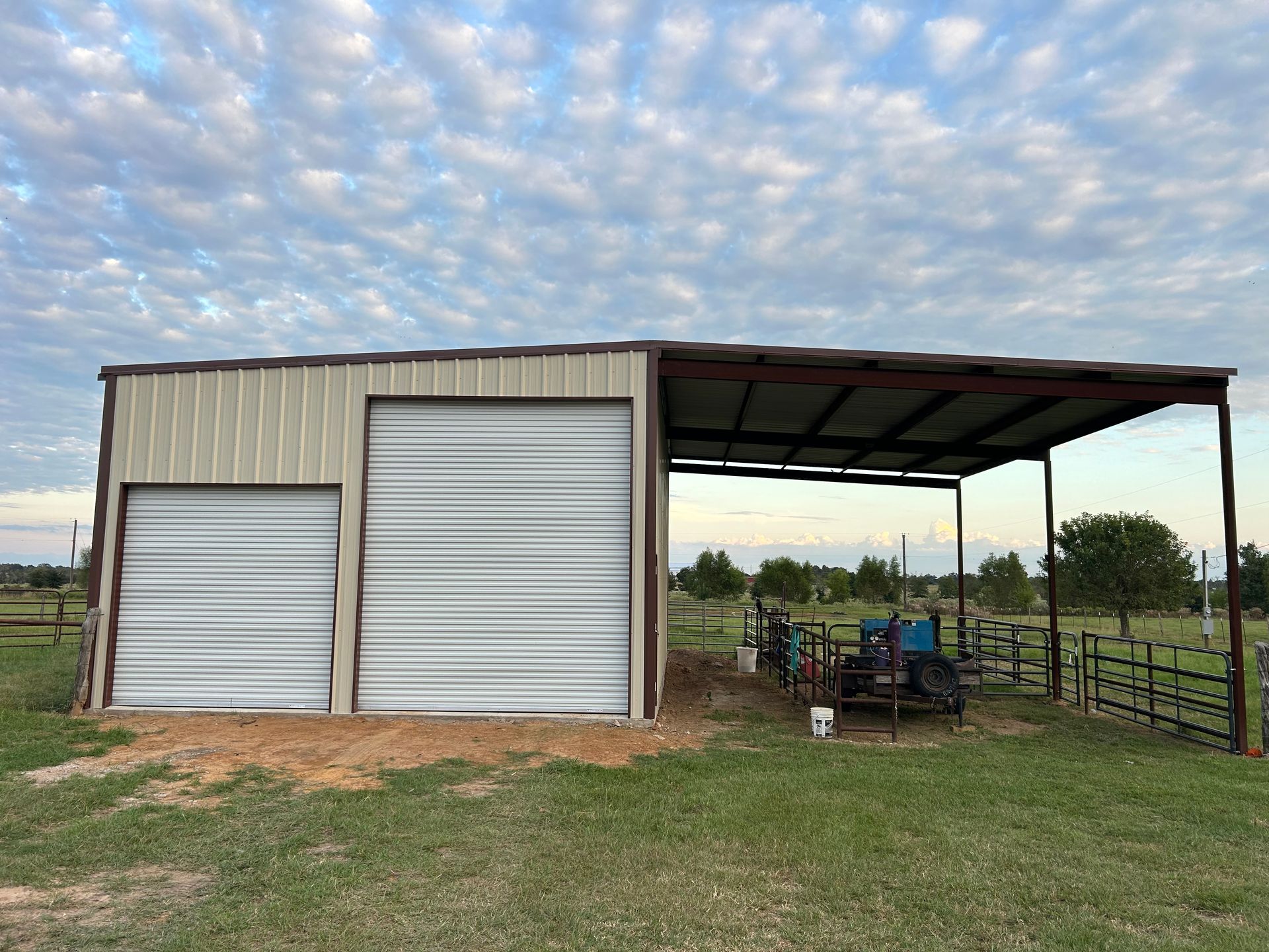 Metal shed with two garage doors, an open awning, and farm equipment outside.