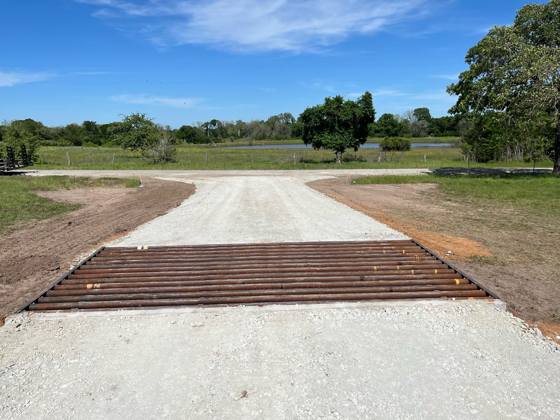 Gravel road with a cattle guard leading into a grassy area, under a bright blue sky.