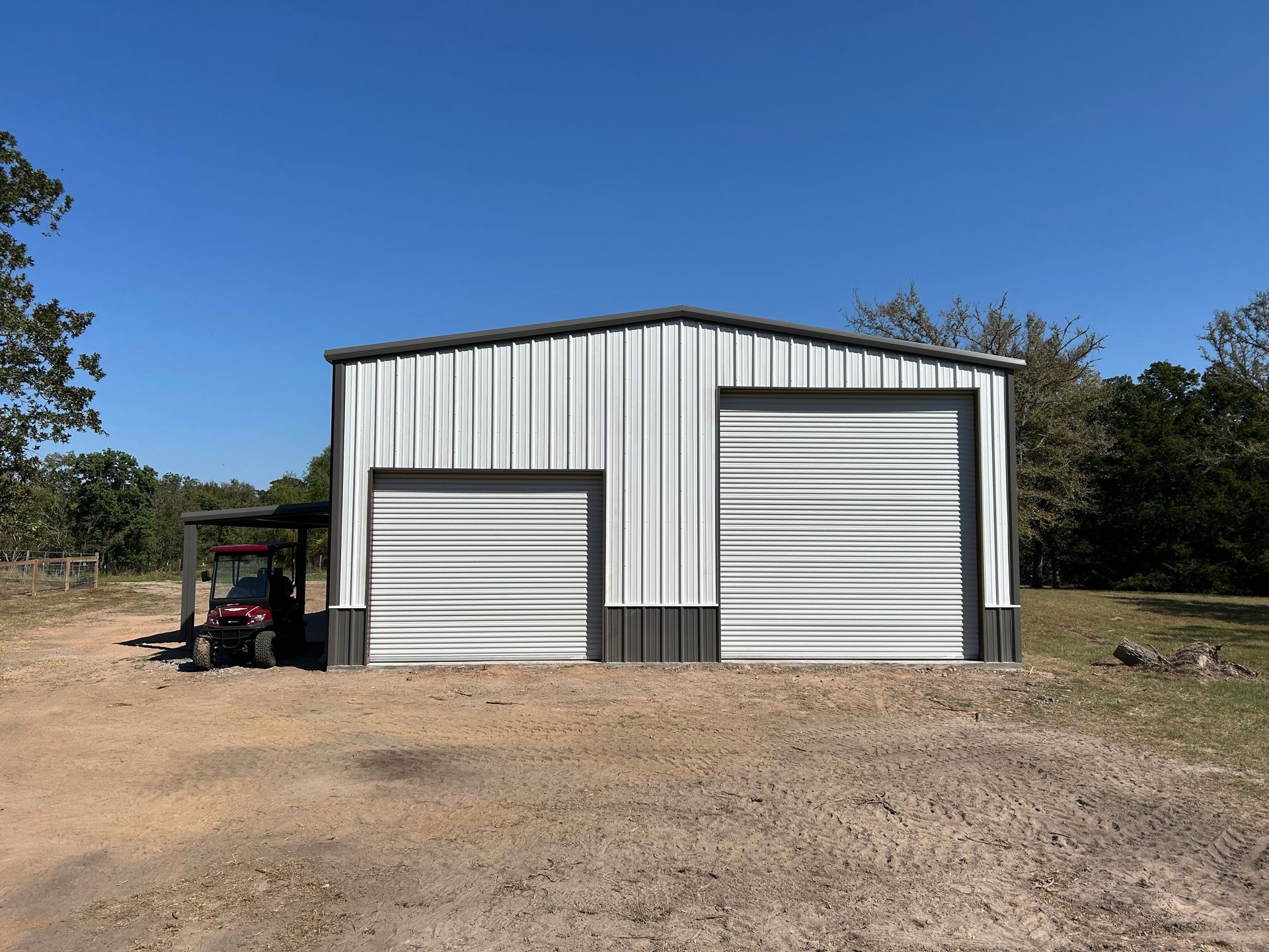 Metal building with two garage doors, tan ground, blue sky. A red vehicle is parked under a carport.