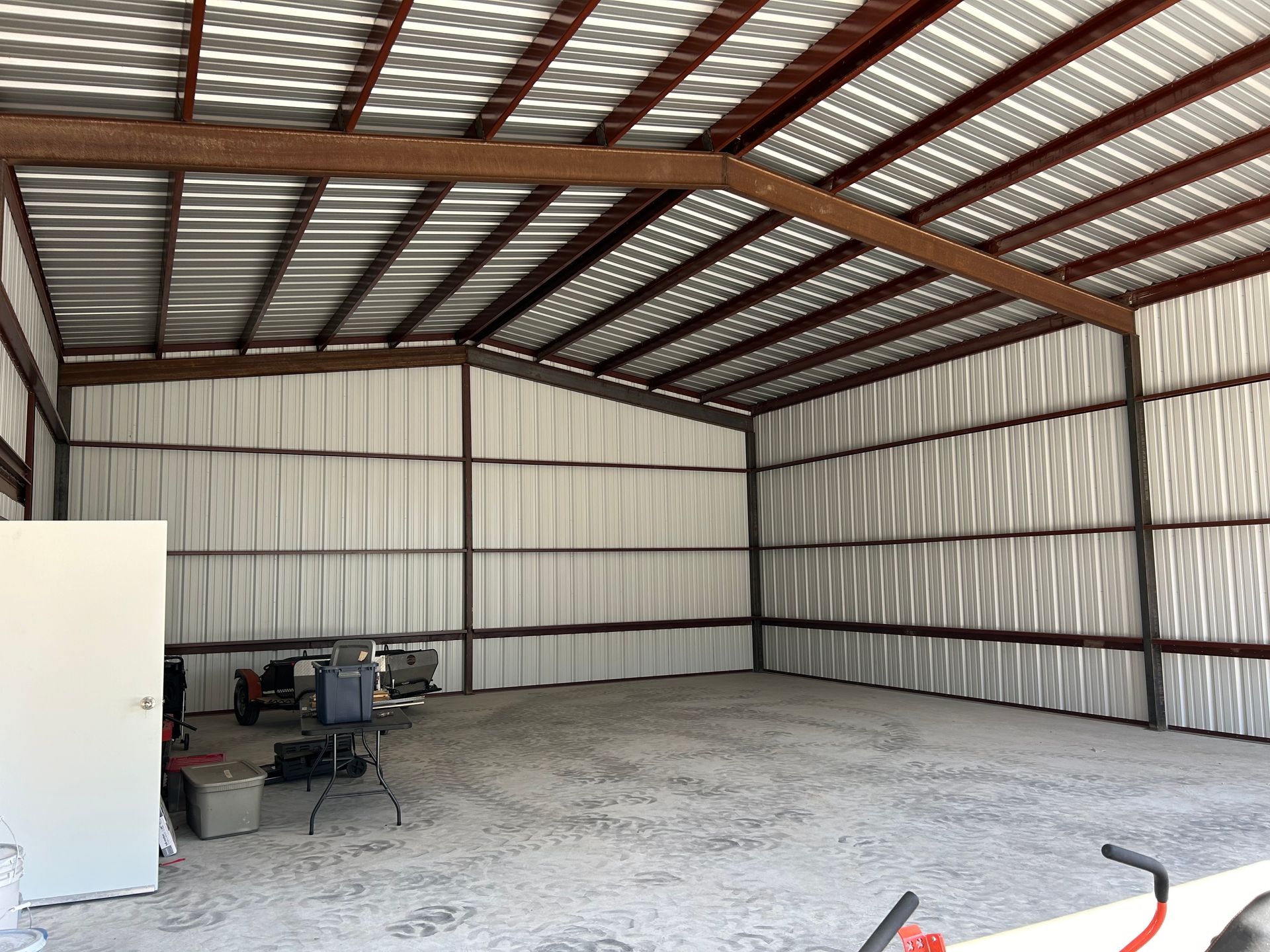 Interior of a metal shed with a concrete floor, brown beams, and tools against a wall.