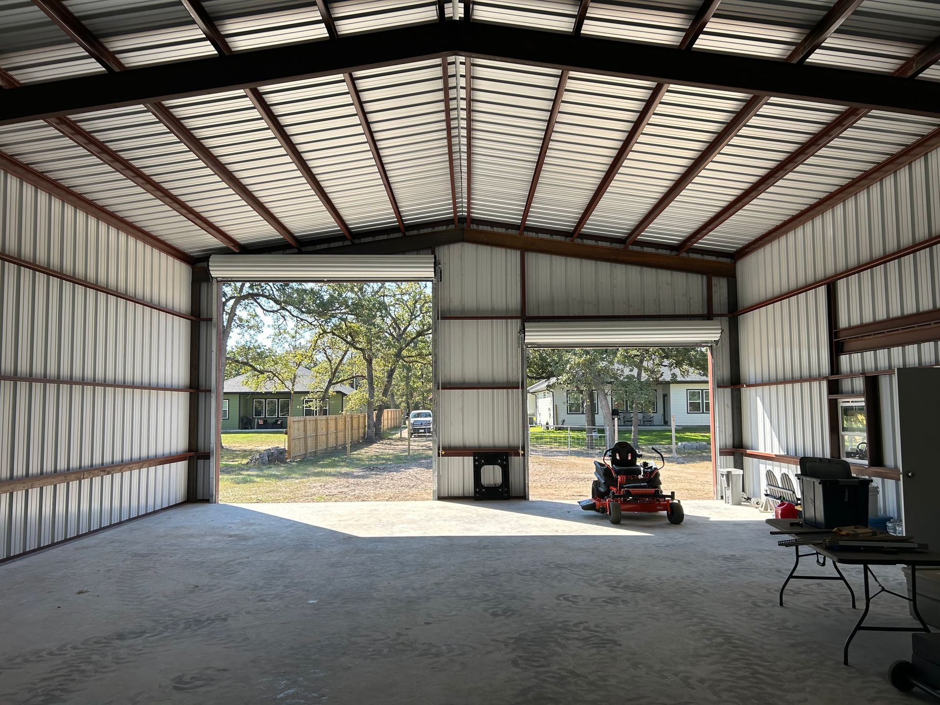 Inside a metal building, two garage doors open to a grassy yard with trees, a riding mower inside.