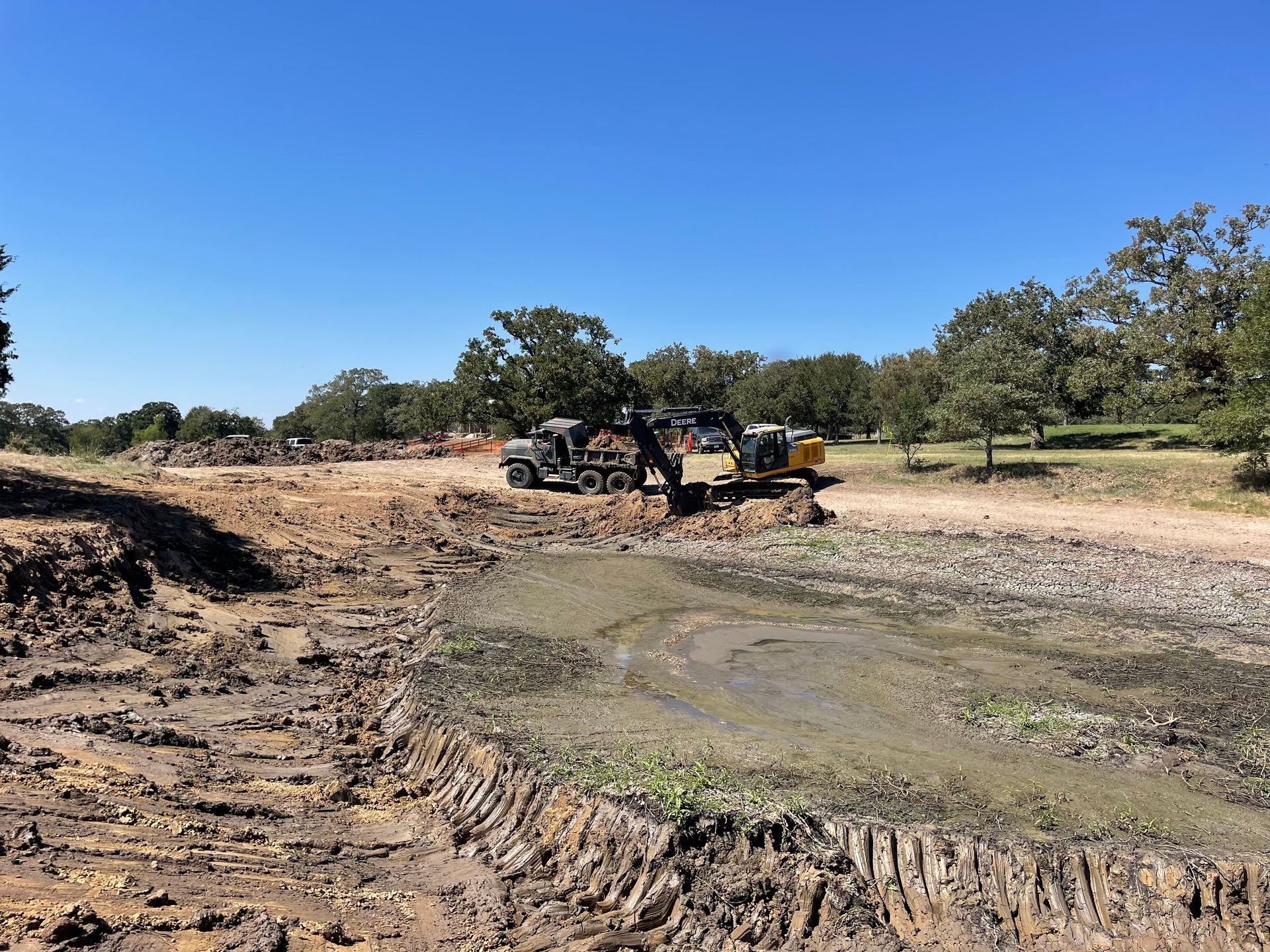 Construction site with heavy machinery, muddy ground, and trees under a blue sky.