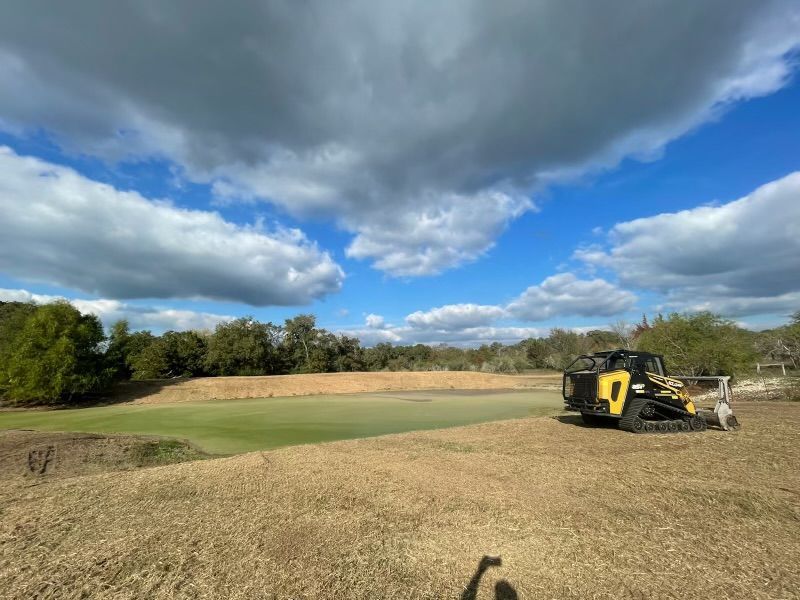 Yellow skid steer clearing dry grass near a green, under a partly cloudy blue sky.