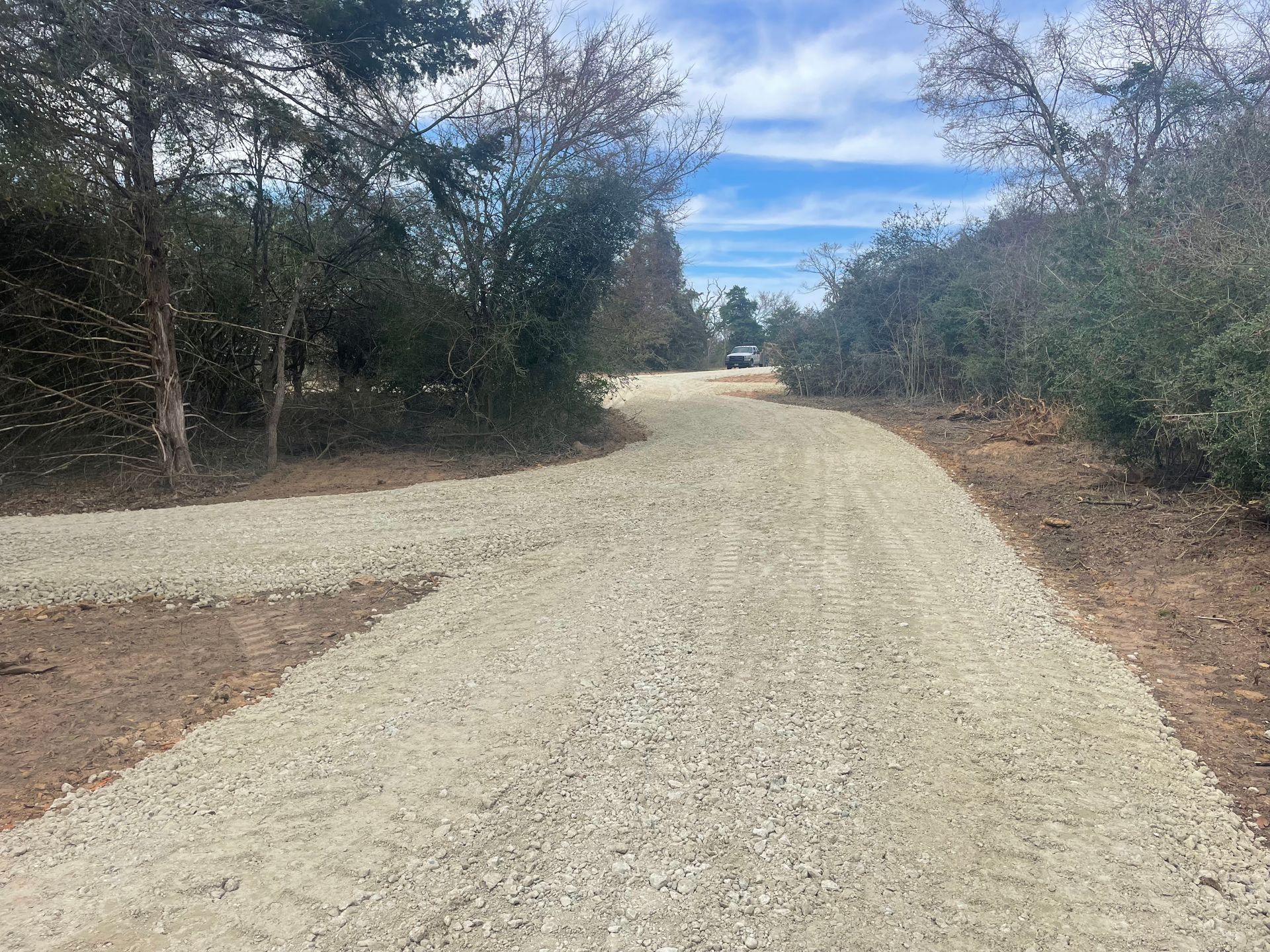 Gravel driveway curving through a wooded area with trees lining both sides under a blue sky.