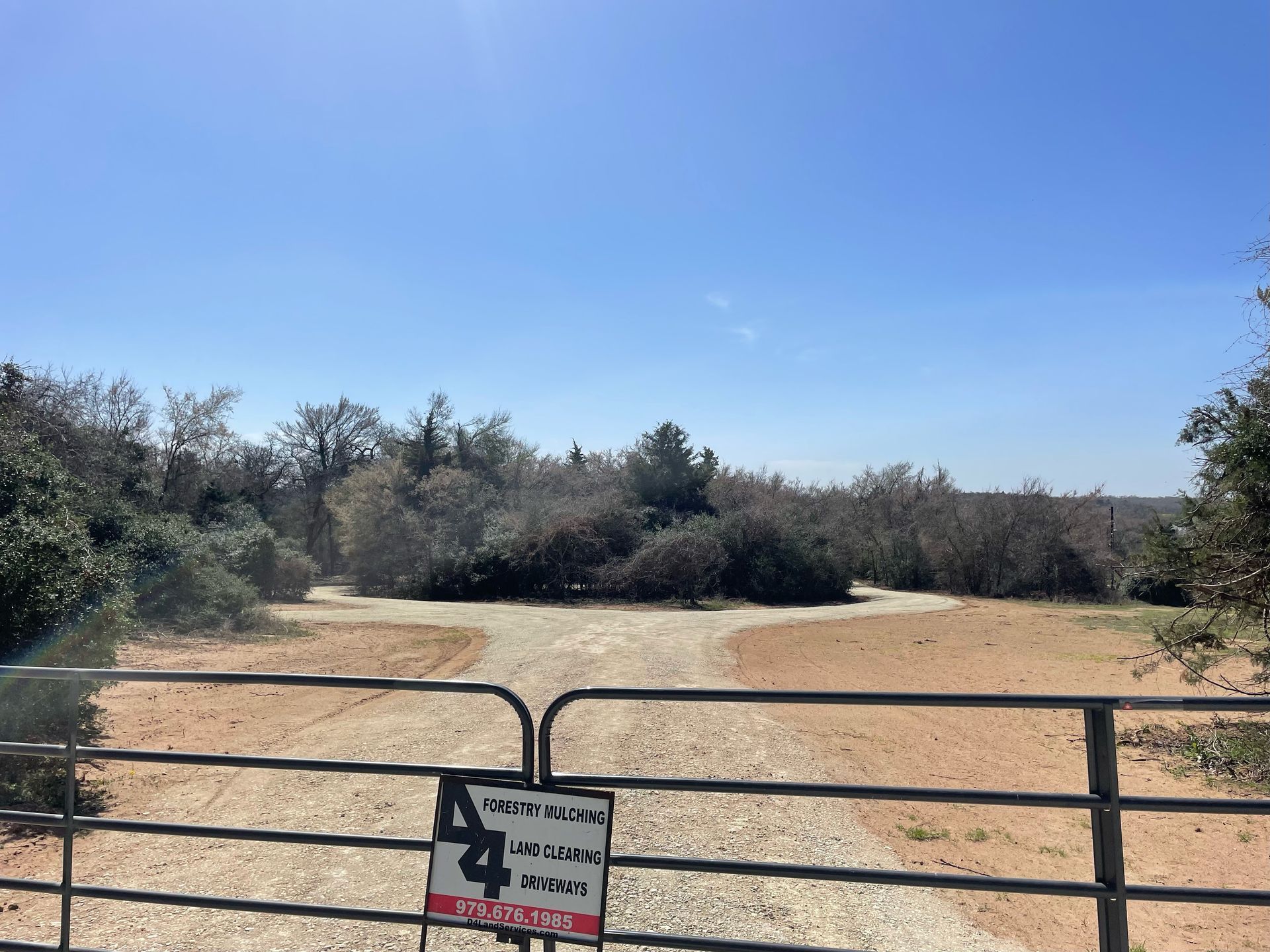 Gate blocking three dirt paths leading into a sunny field with scrub brush and trees.