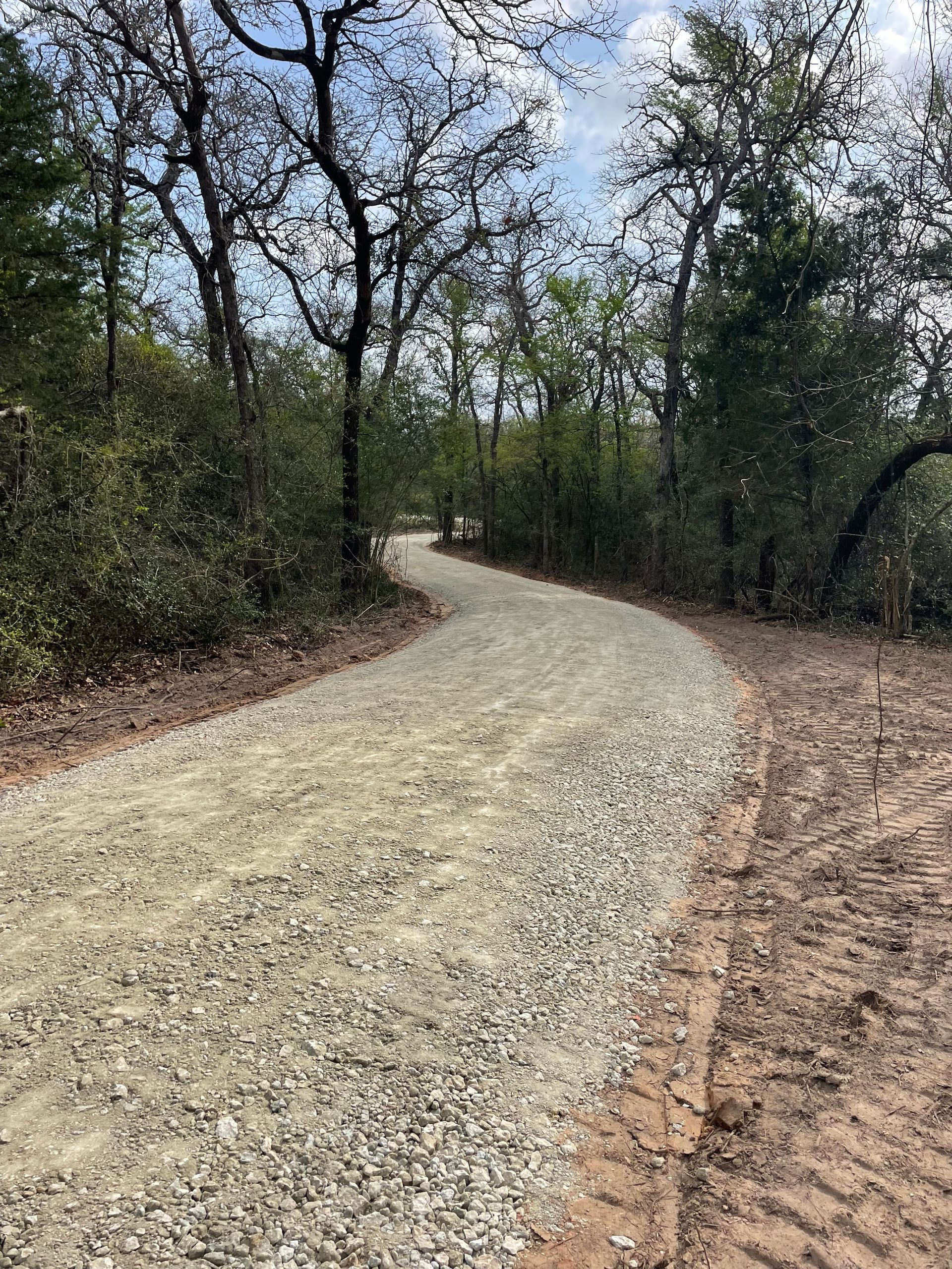 Gravel path winding through a wooded area with trees and bushes under a cloudy sky.