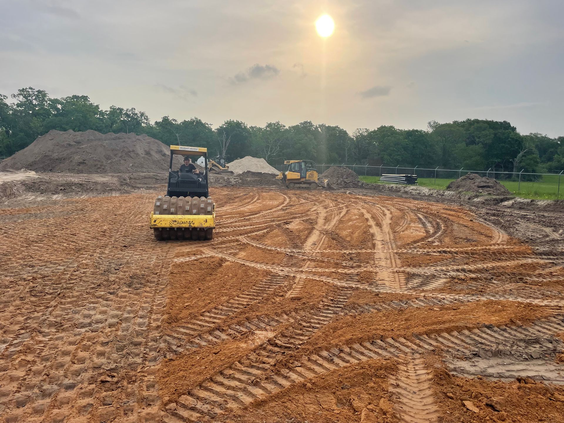 A construction site with a roller compactor flattening dirt. A bulldozer and piles of dirt are in the background.