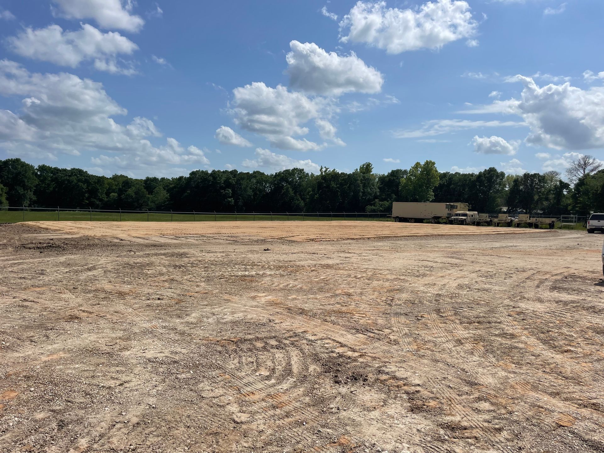 Cleared, flat, dirt lot under a blue sky with fluffy clouds and a line of trees in the distance.