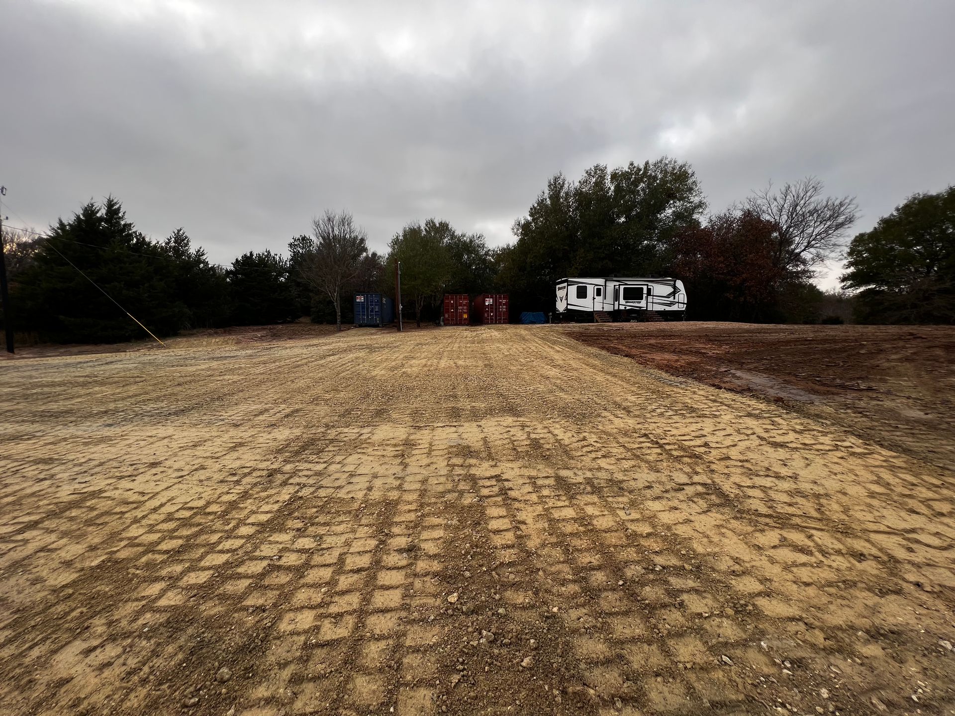 Dirt road with tire tracks leading to trailers, containers, trees, under an overcast sky.
