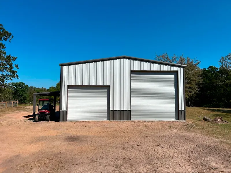 White metal building with two garage doors, tractor under a lean-to, blue sky.