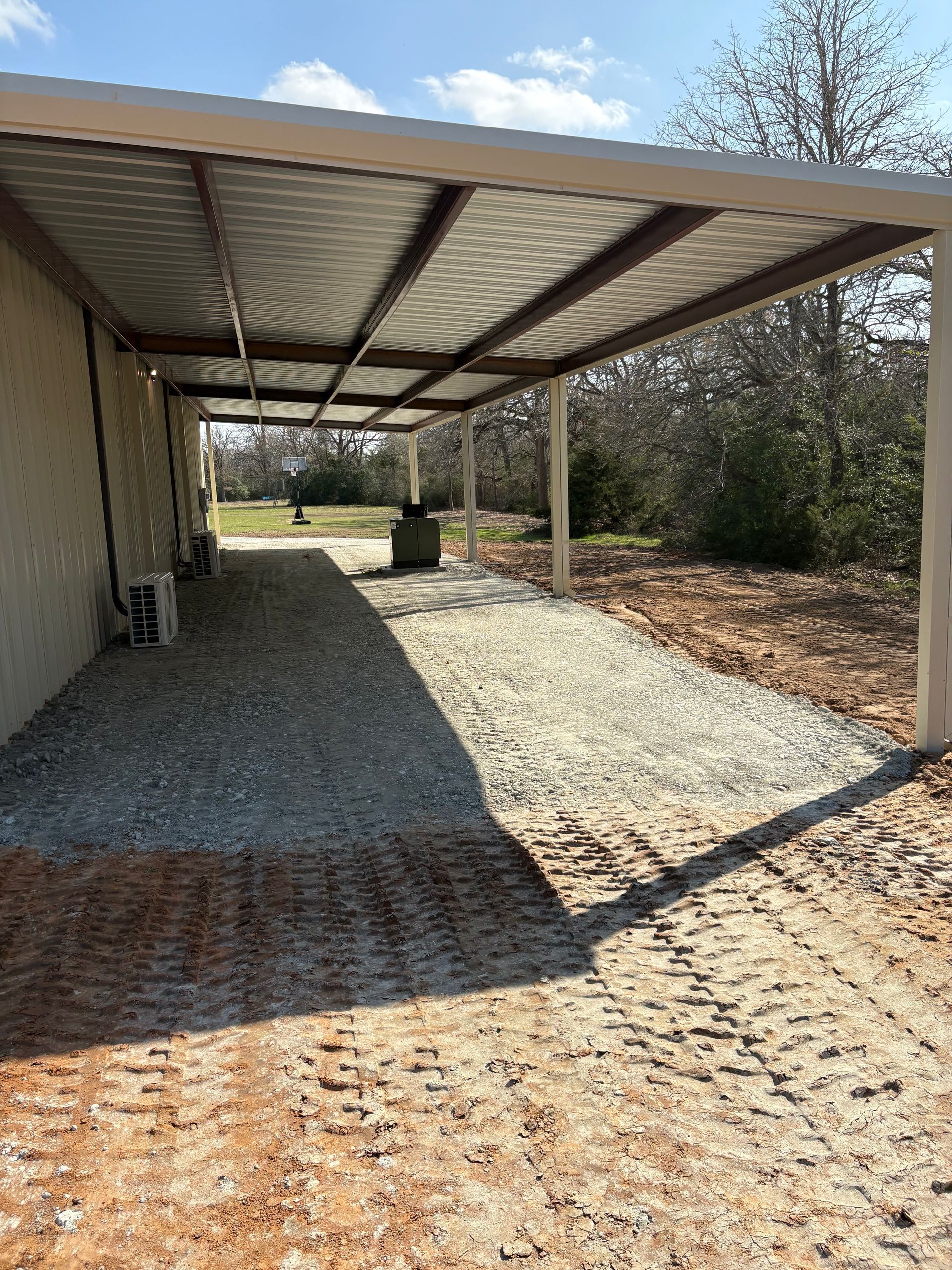 A covered gravel driveway next to a building with a metal roof on a sunny day.