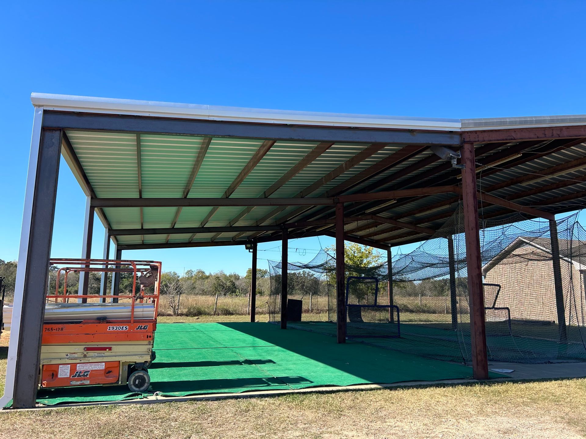 Metal baseball batting cage with green turf under a blue sky.