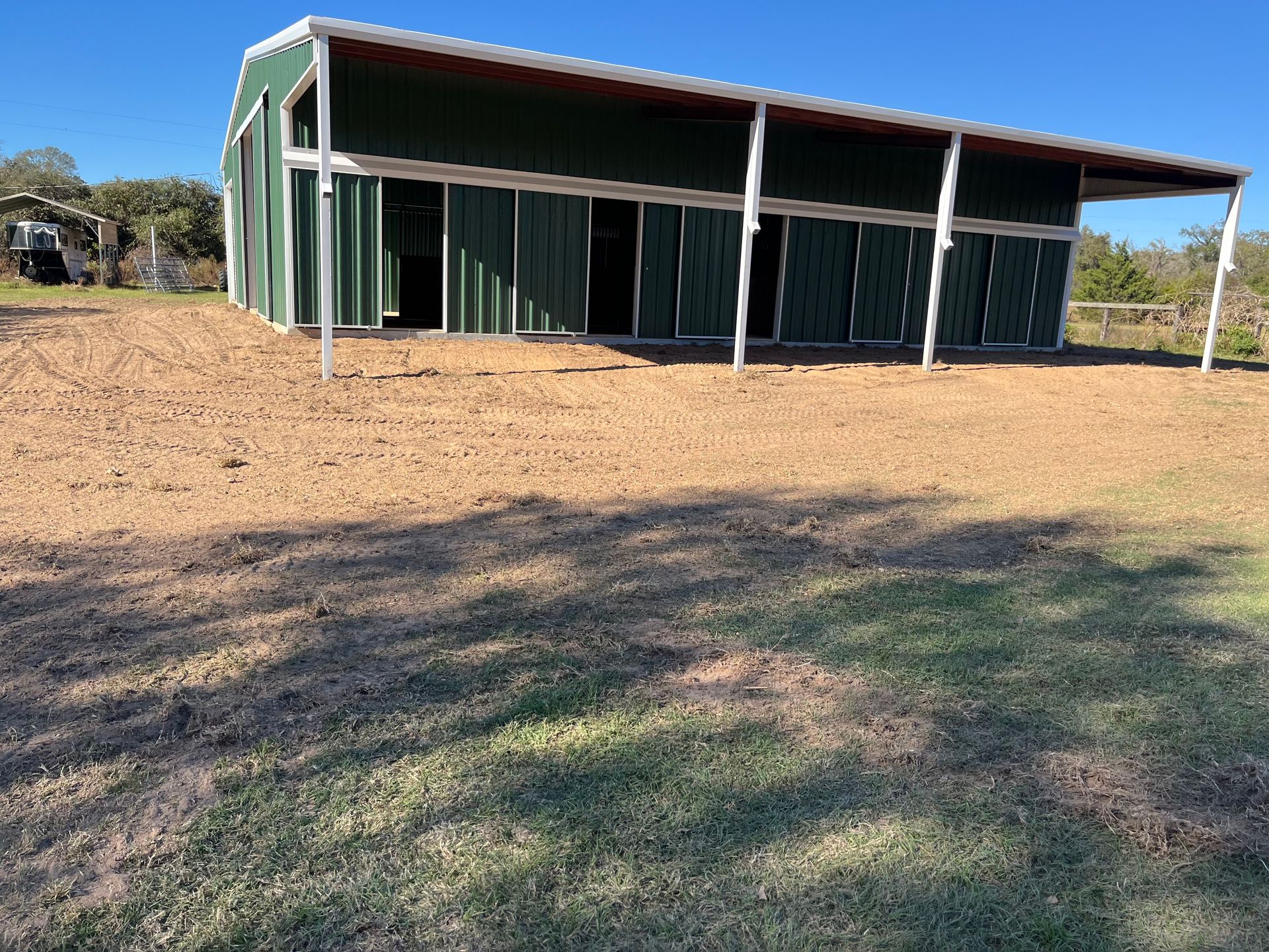 Green barn with white trim and a brown dirt lot on a sunny day.