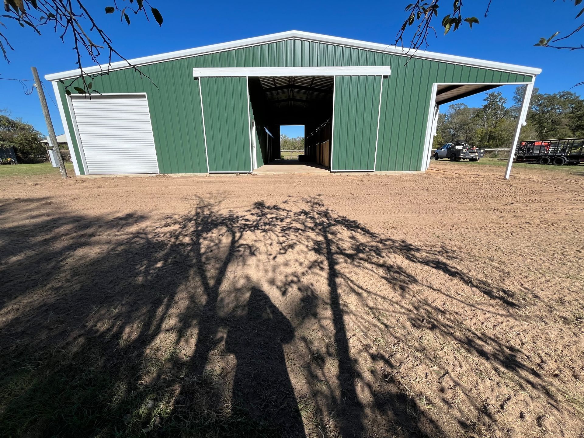 Green metal barn with open center, white trim, on dirt; tree shadow in foreground.