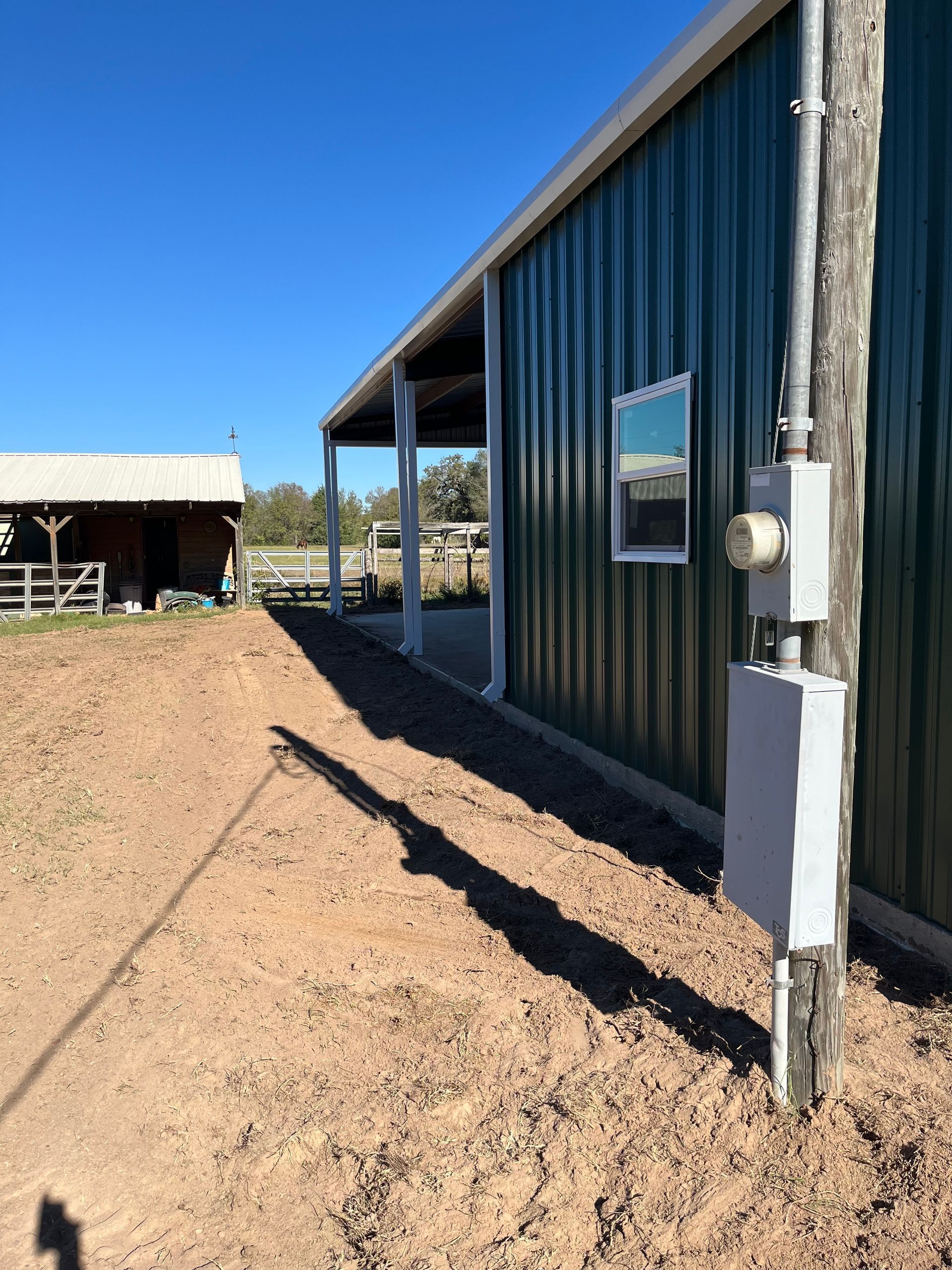 Green metal building with a porch and utility pole on a dirt path, sunny day.