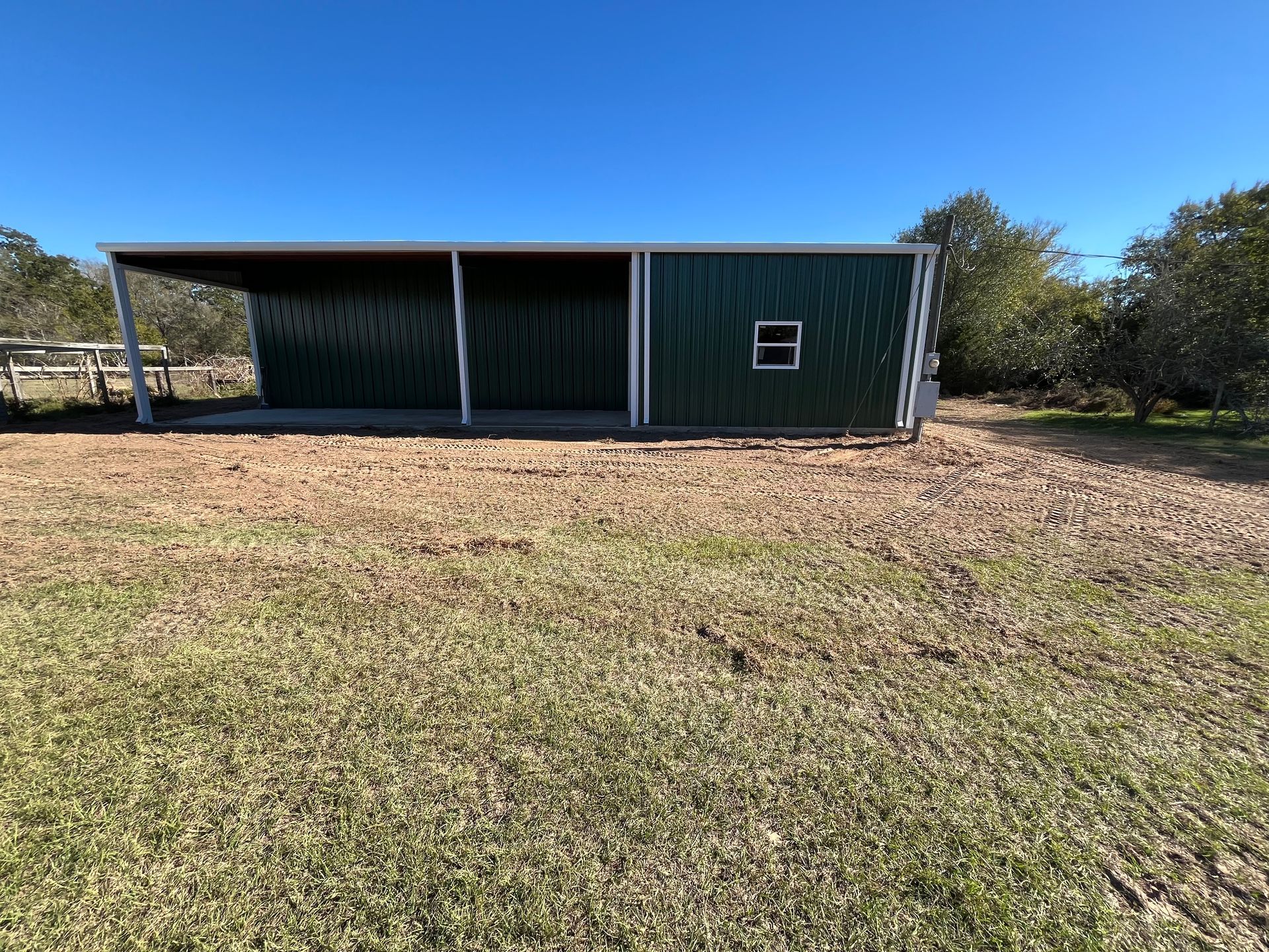 Green metal shed with a covered area, on a grassy field, under a clear blue sky.