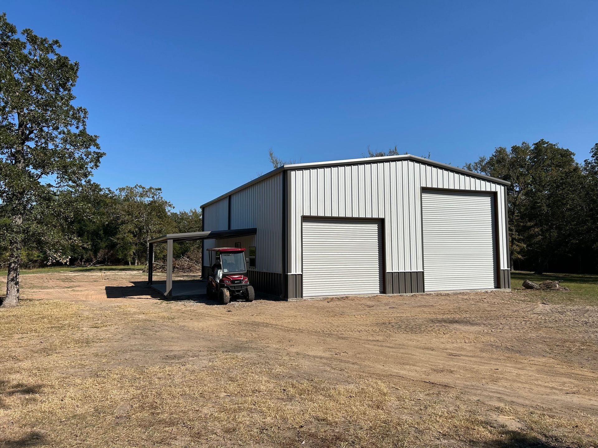 A metal workshop with two garage doors and a side awning; a red utility vehicle is parked under the awning.