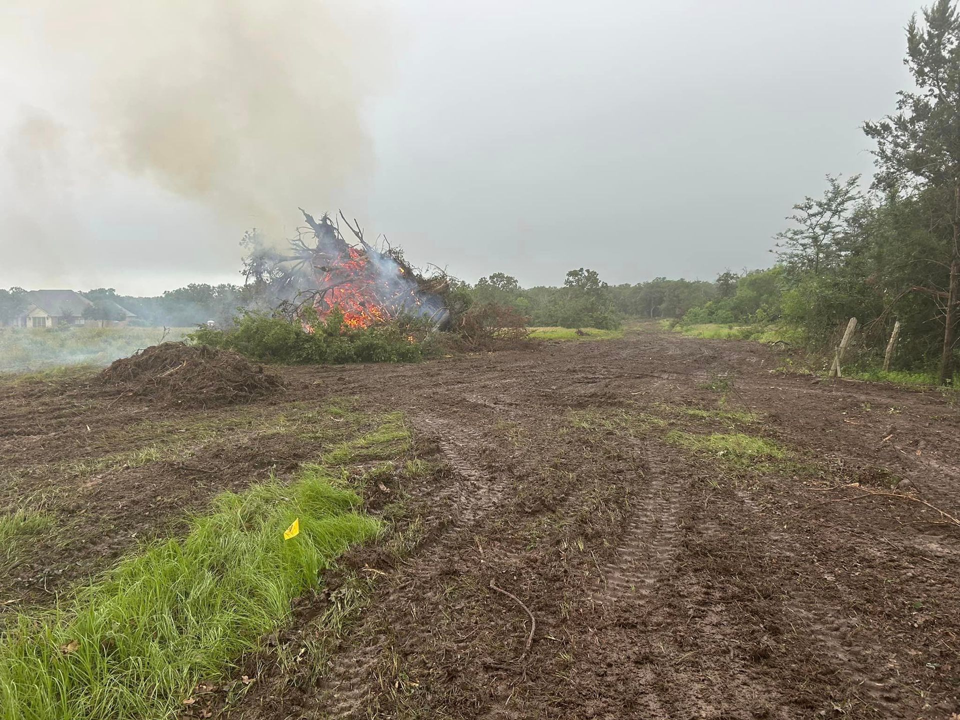 A large brush pile burns, emitting smoke, in a muddy field; trees in background.