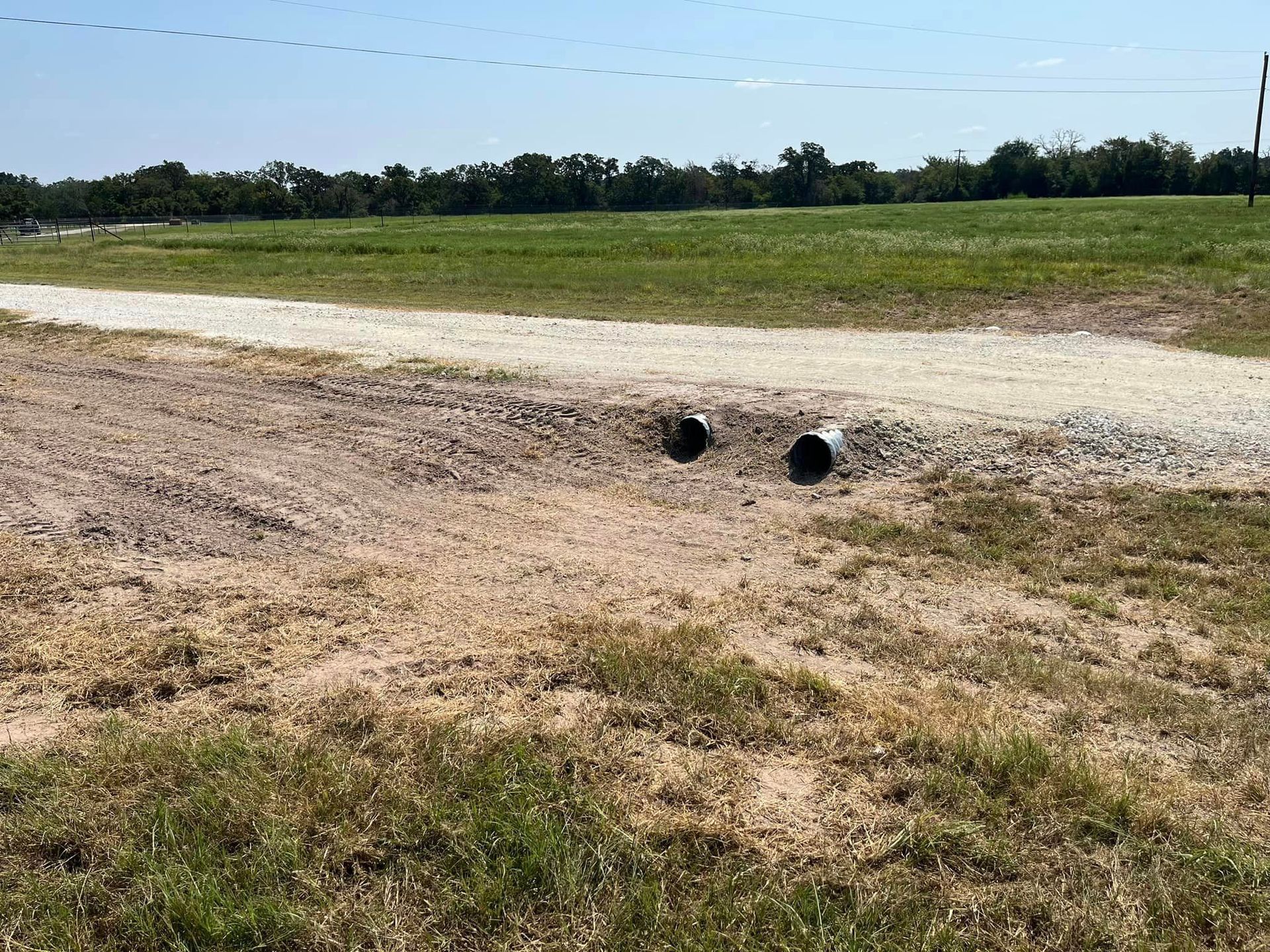 Two drainage pipes under a gravel road in a grassy field.