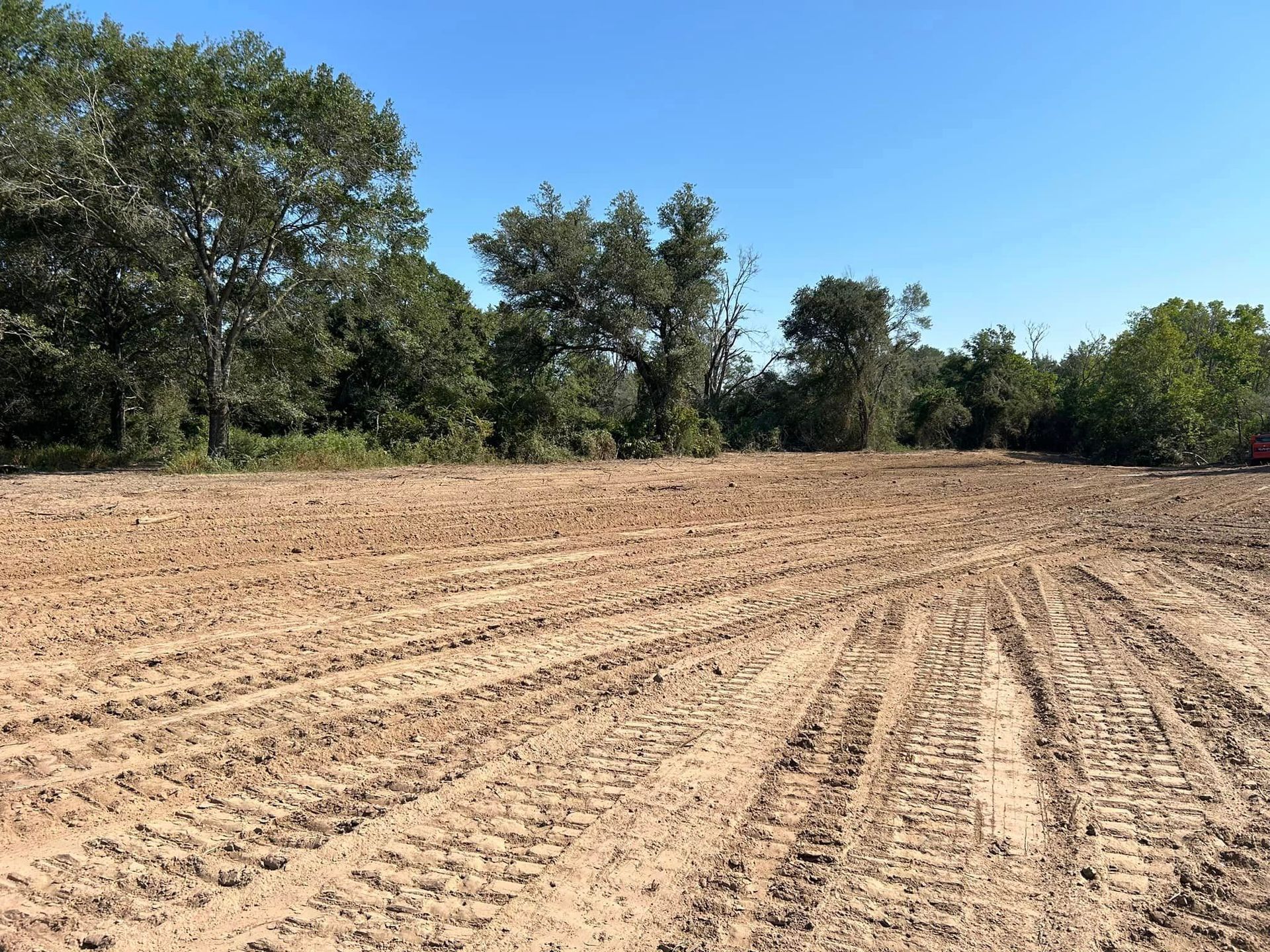 Tilled dirt field with tire tracks, trees in background, and a clear blue sky.