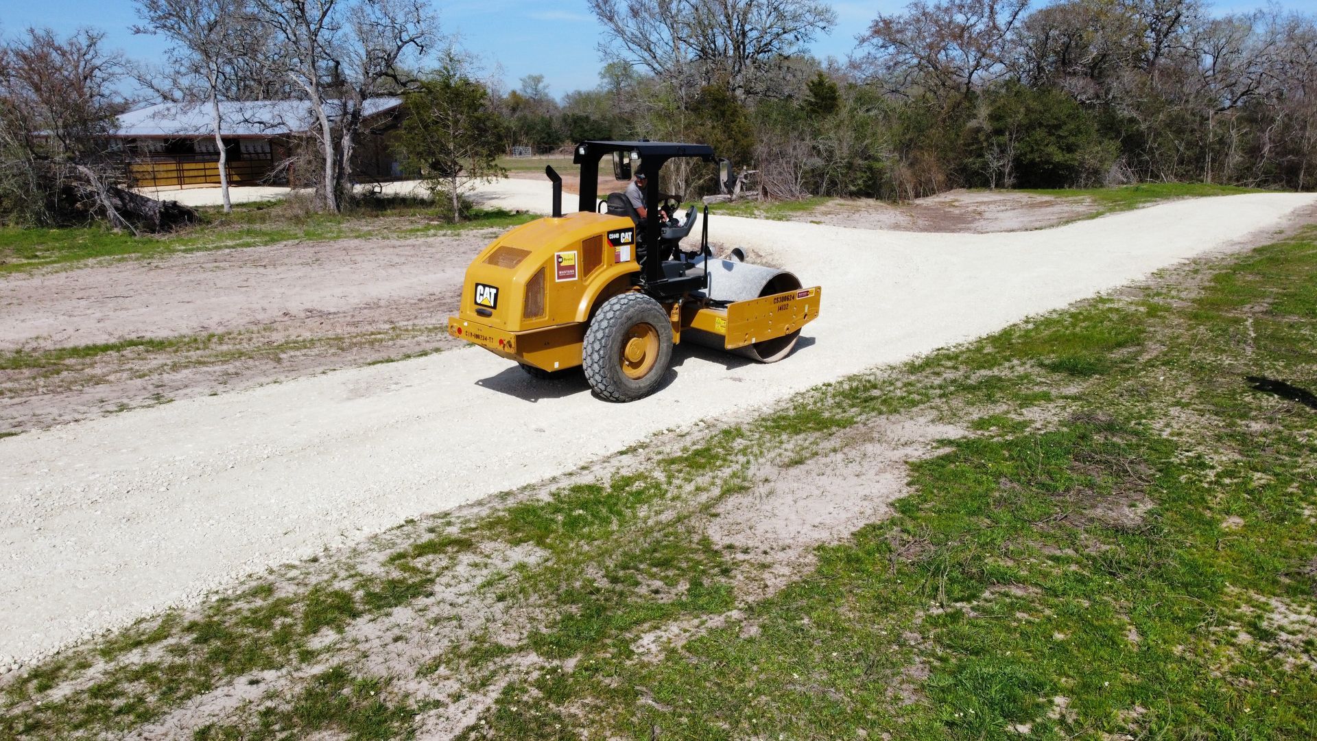 Yellow road roller compacting gravel road. Rural setting with green grass and trees.