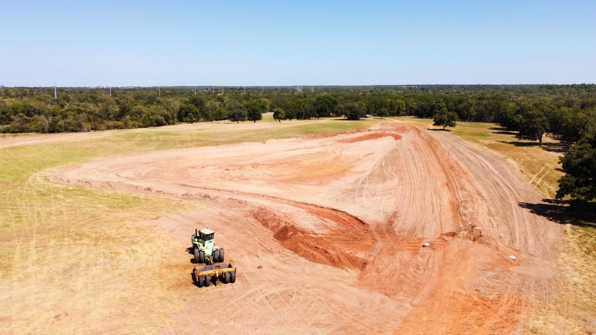 Tractor plowing a field of red soil, surrounded by green grass and trees under a blue sky.