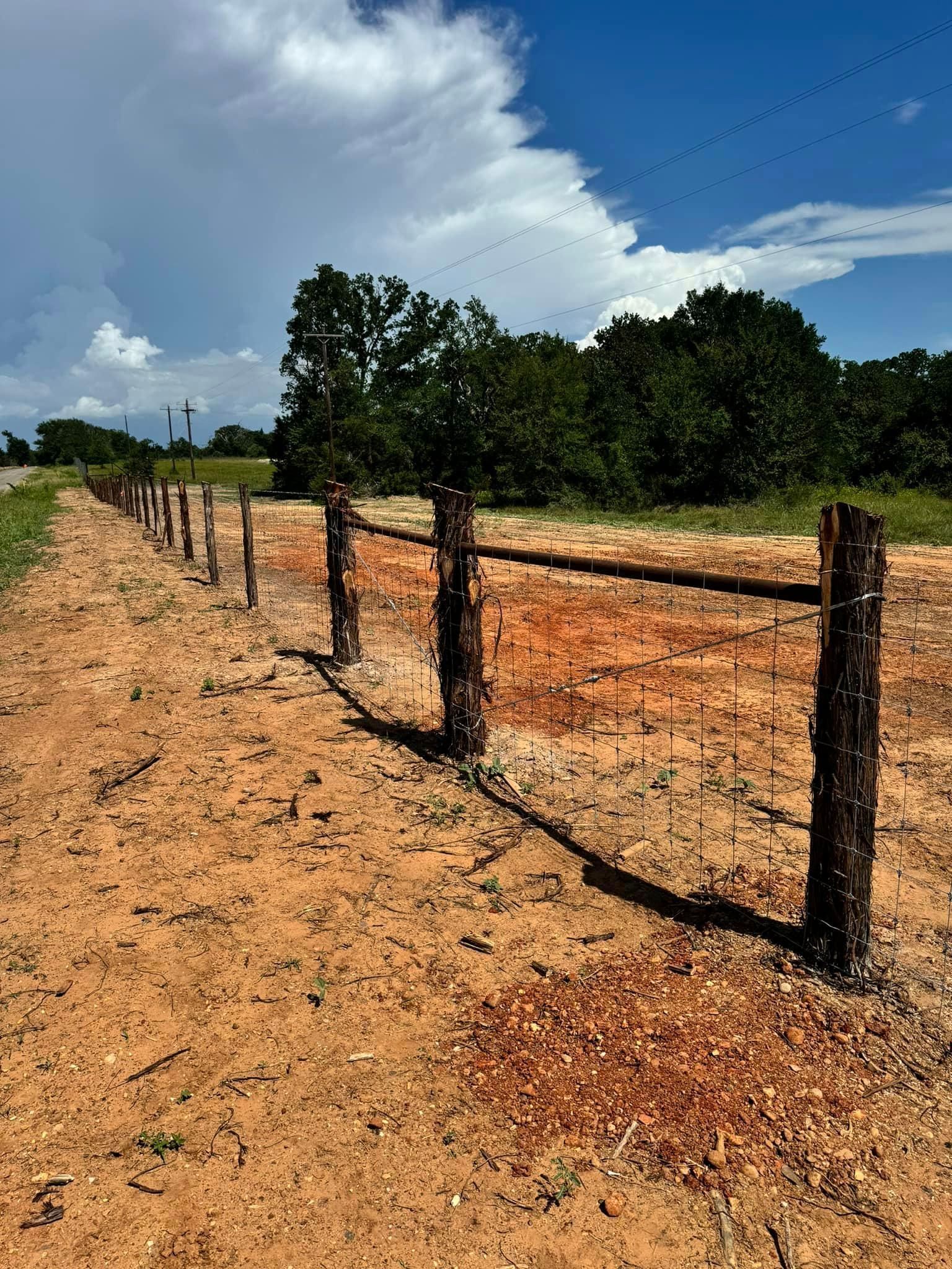 Fence in a dirt field, with trees in the background under a cloudy sky.