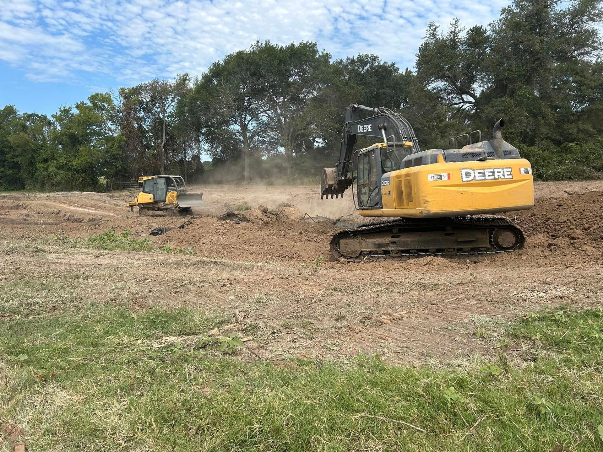 Two John Deere construction machines working on a dirt field, trees in the background.