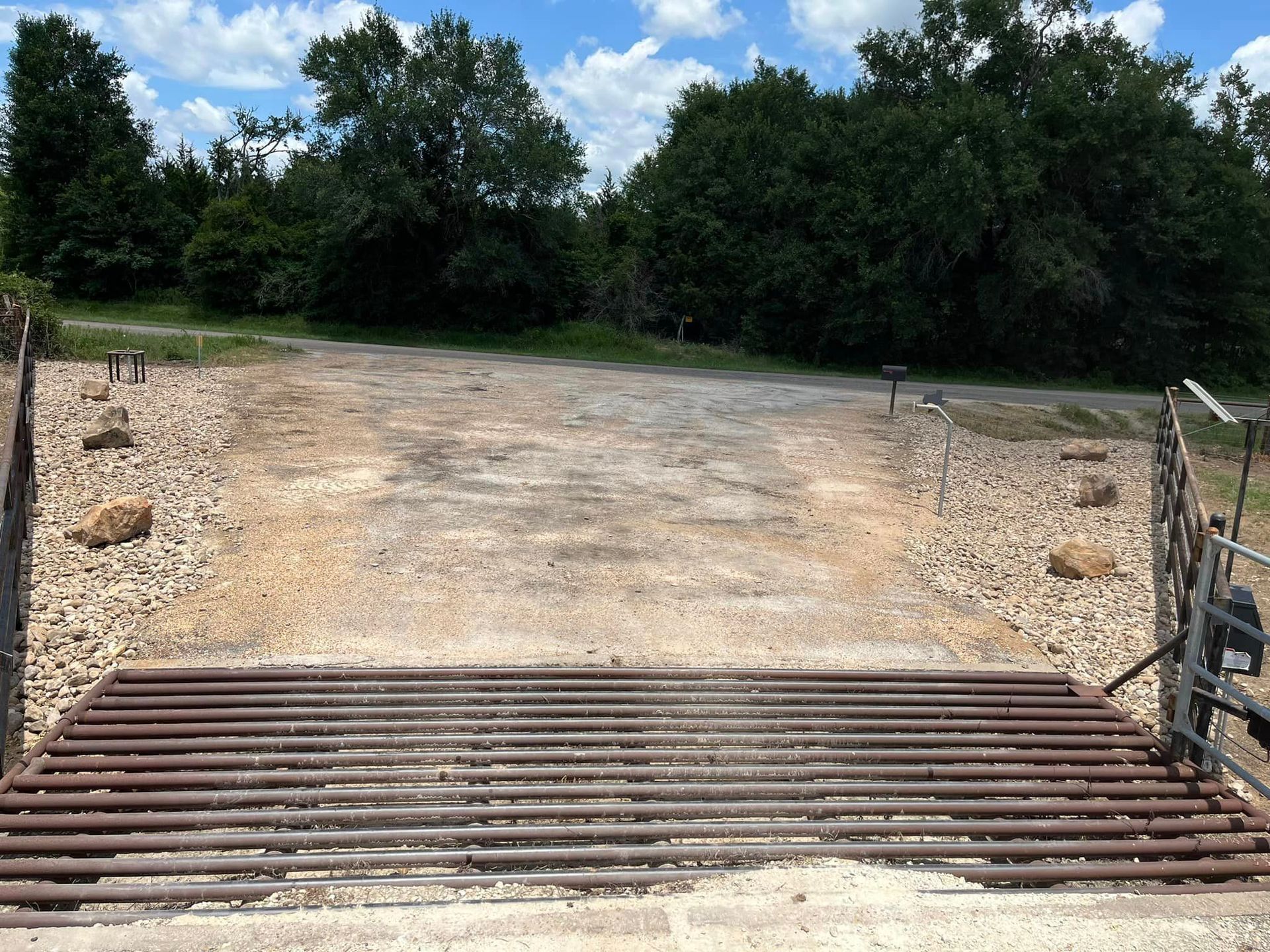 Cattle guard in a dirt road leading to a wooded area under a blue sky.
