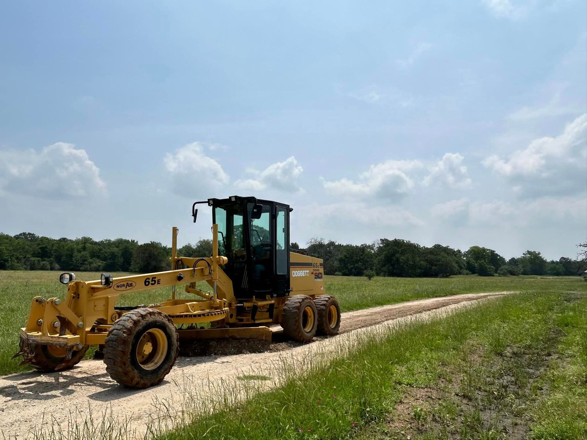 Yellow grader on a gravel road through a field under a bright, cloudy sky.