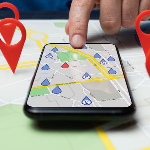 Hand pointing at a smartphone map displaying location pins; red and blue pins, map is on a table.