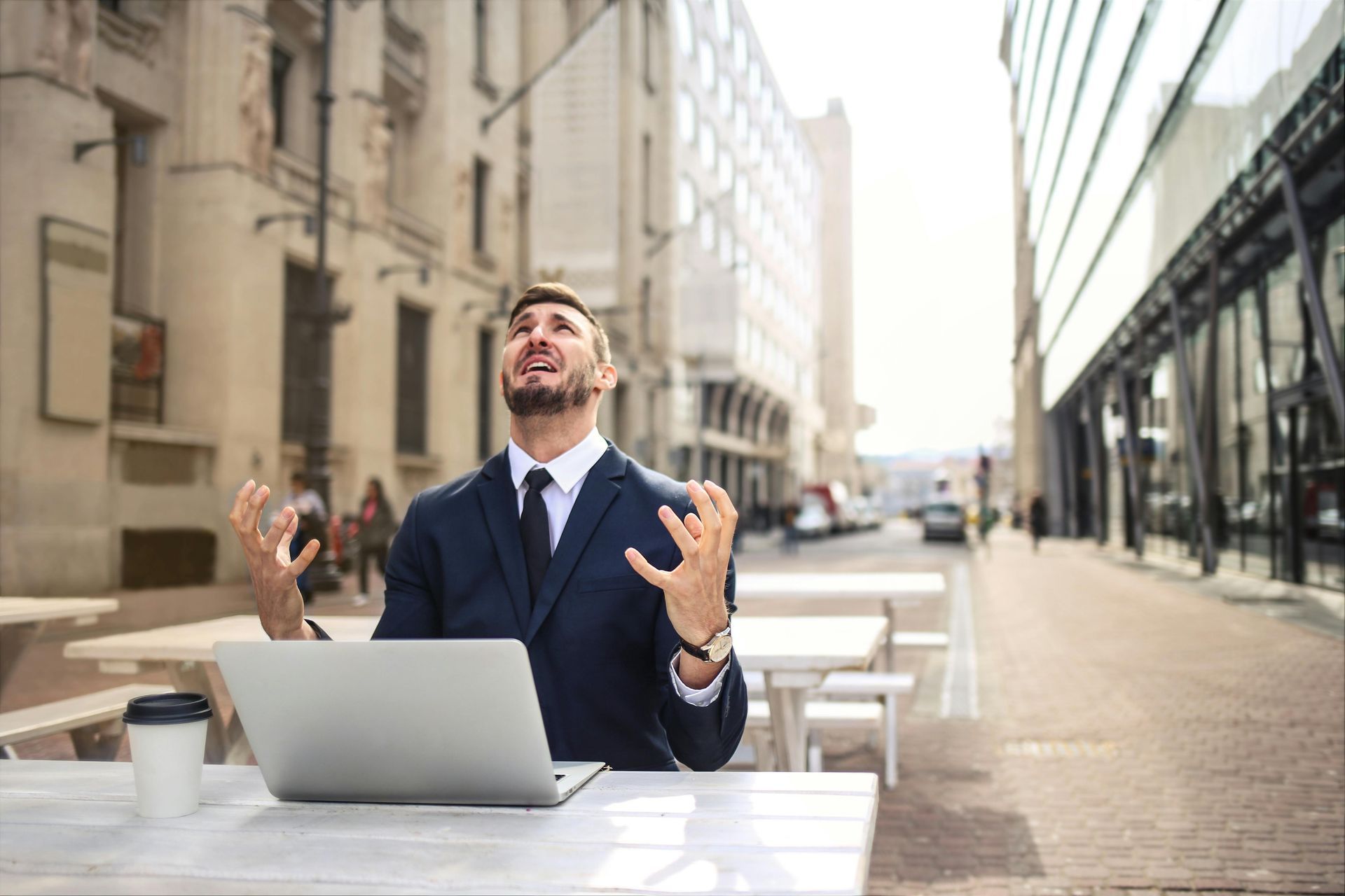 Businessman in suit looking frustrated, arms raised, sitting at table with laptop and coffee outdoors.