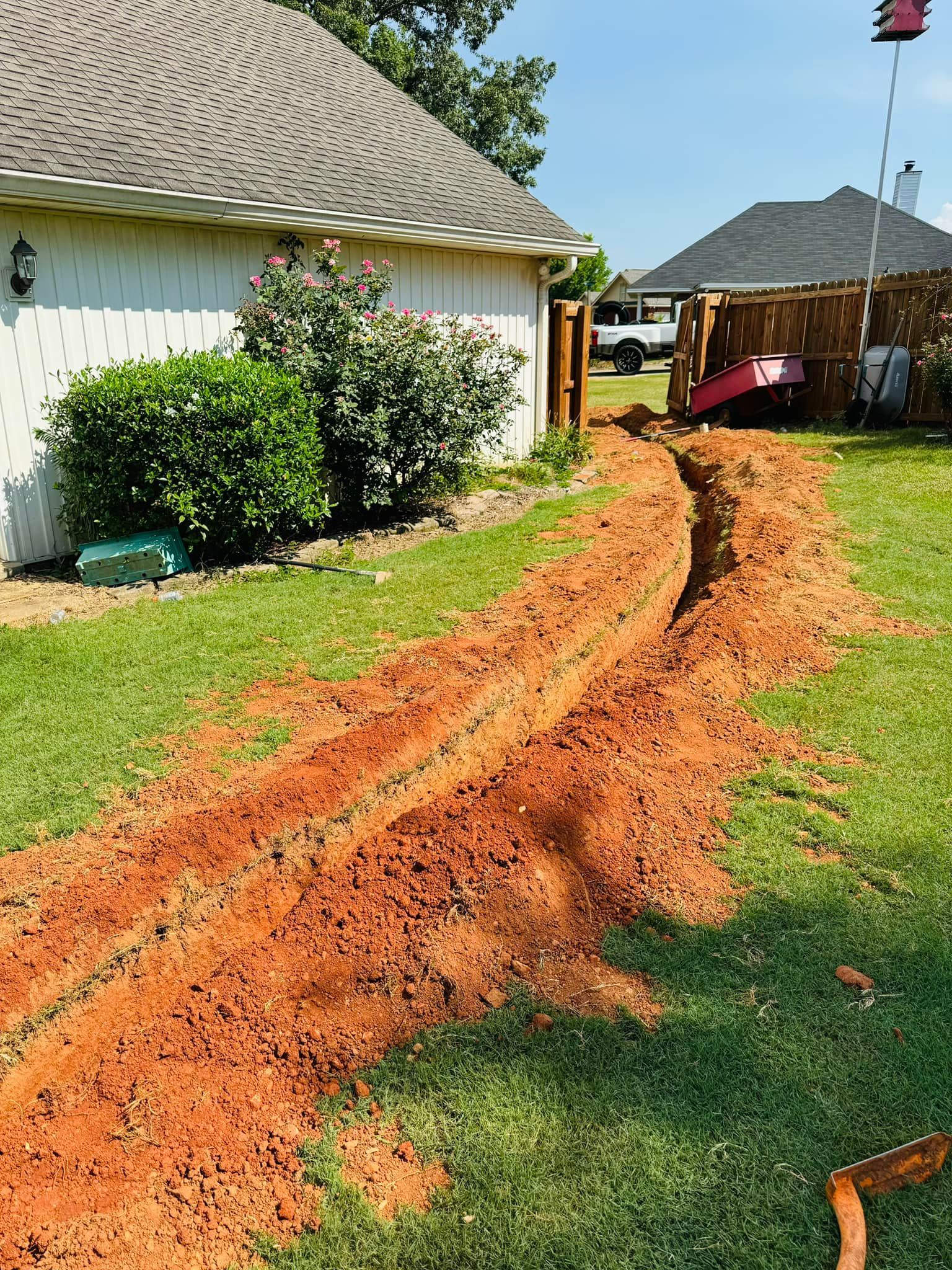 Trench dug in a grassy yard, leading to a fence. The trench is filled with red soil, next to a house.