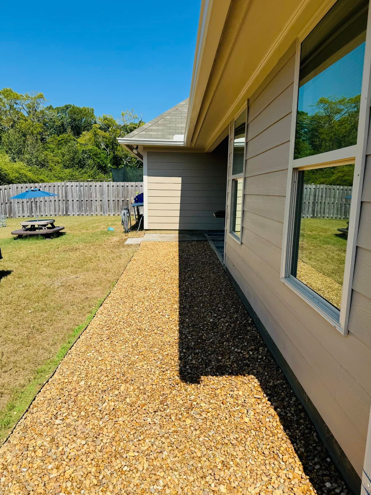Gravel pathway alongside a beige house with reflective windows, leading to a grassy backyard with a picnic table and fence.