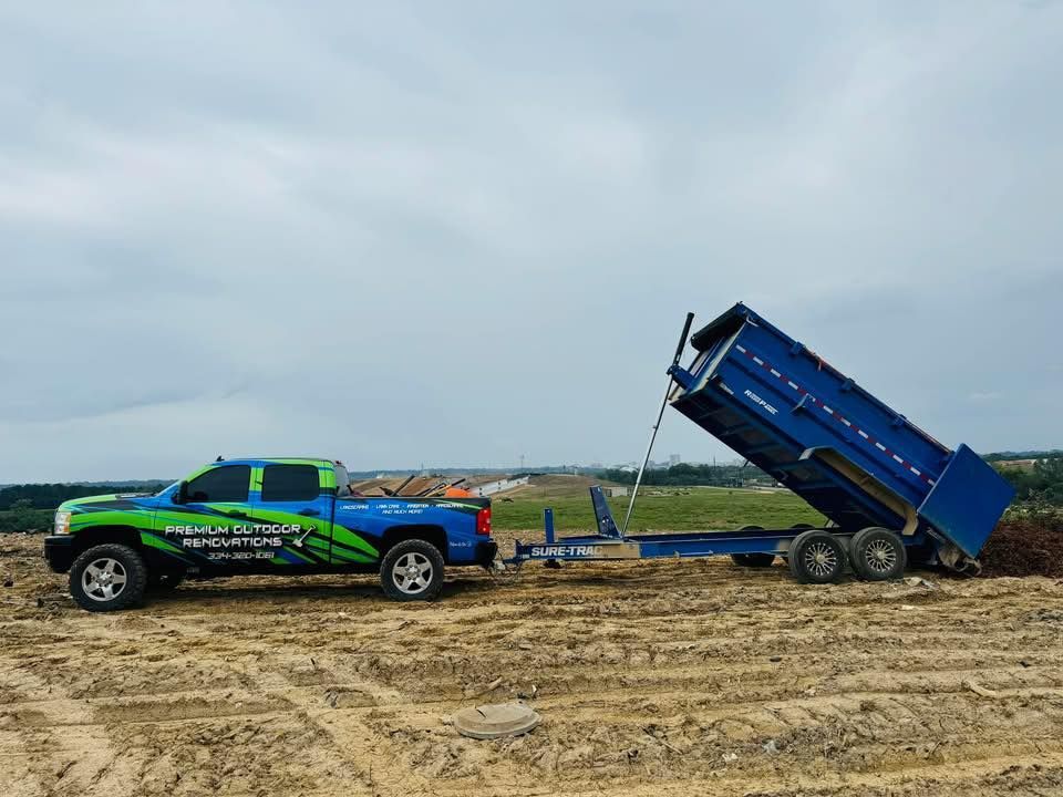 Green and blue truck towing a blue dump trailer, parked on a muddy, outdoor worksite under a cloudy sky.