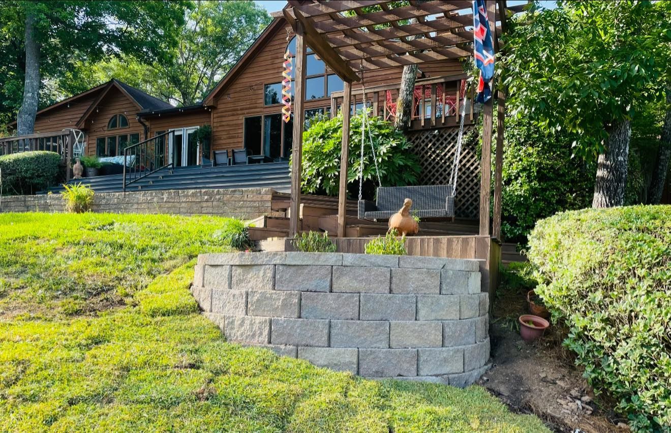 Wooden house with deck overlooking a lake, accessed by a stone staircase. Arbor with swing. Lush green lawn and trees.