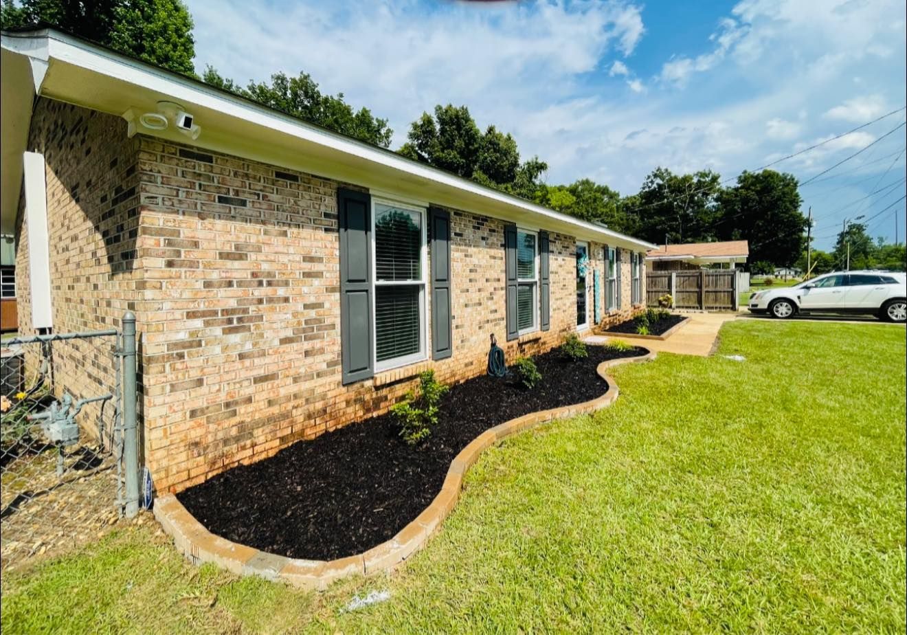 Brick house with black shutters, landscaping, and a car in the driveway.