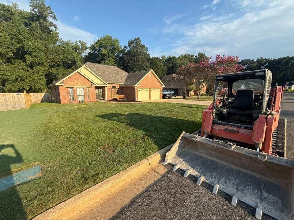Orange skid steer on street near a brick house, pushing material. Green lawn and trees.
